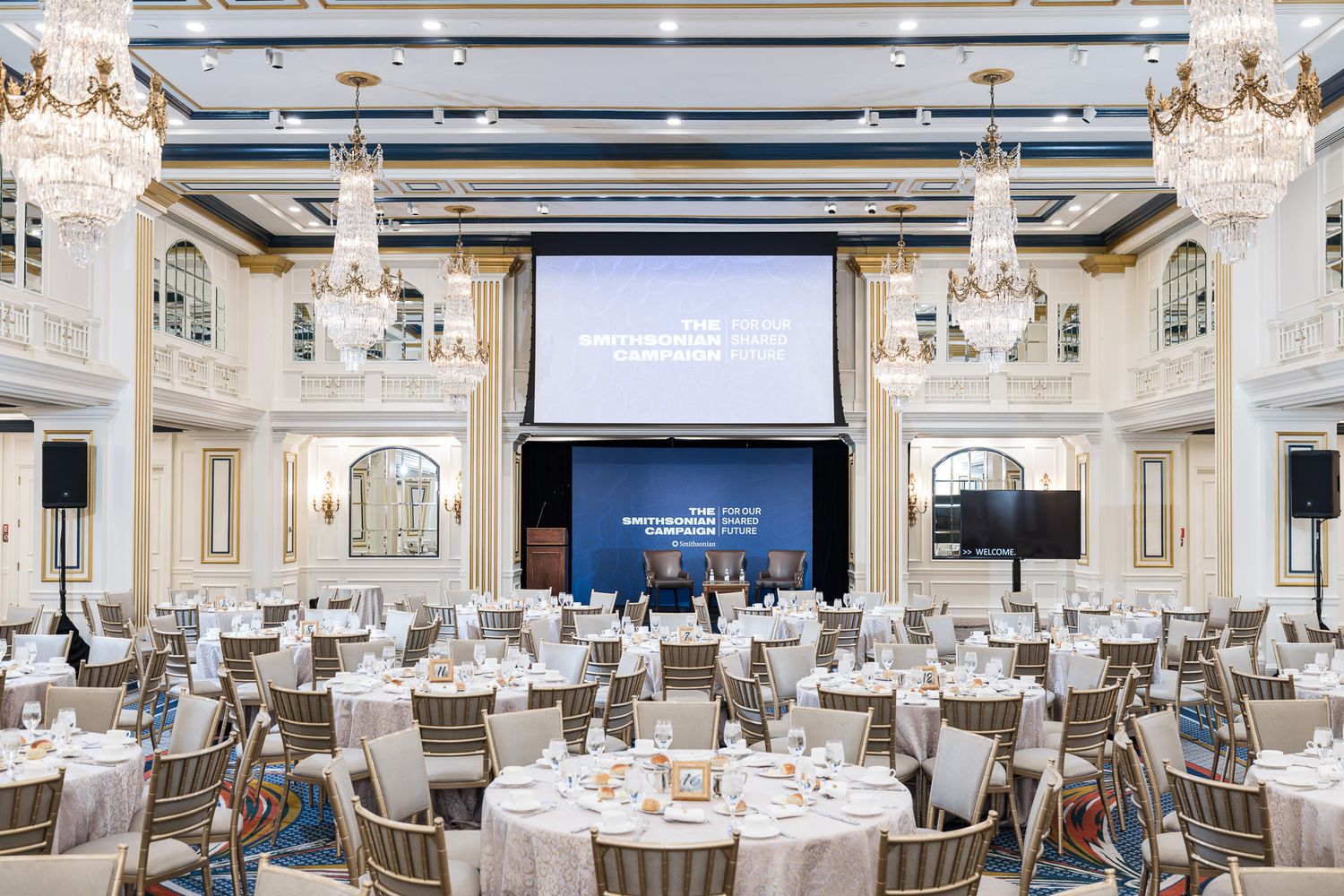 Elegant ballroom with crystal chandeliers, white decor, and round dining tables set for a formal event.