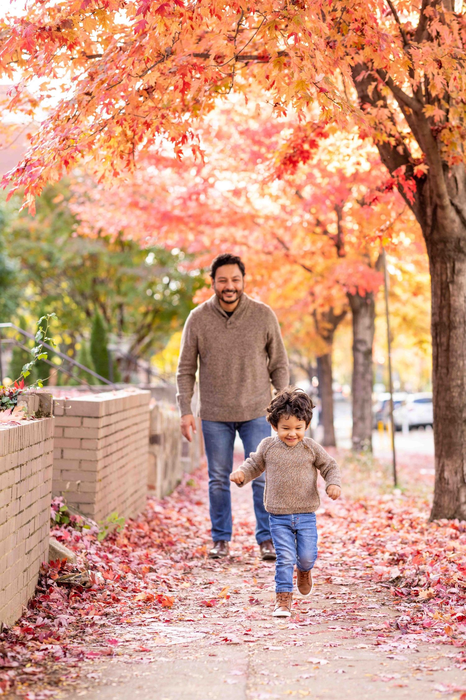 Father and child holding hands, running on a sidewalk surrounded by vibrant red and orange autumn foliage.