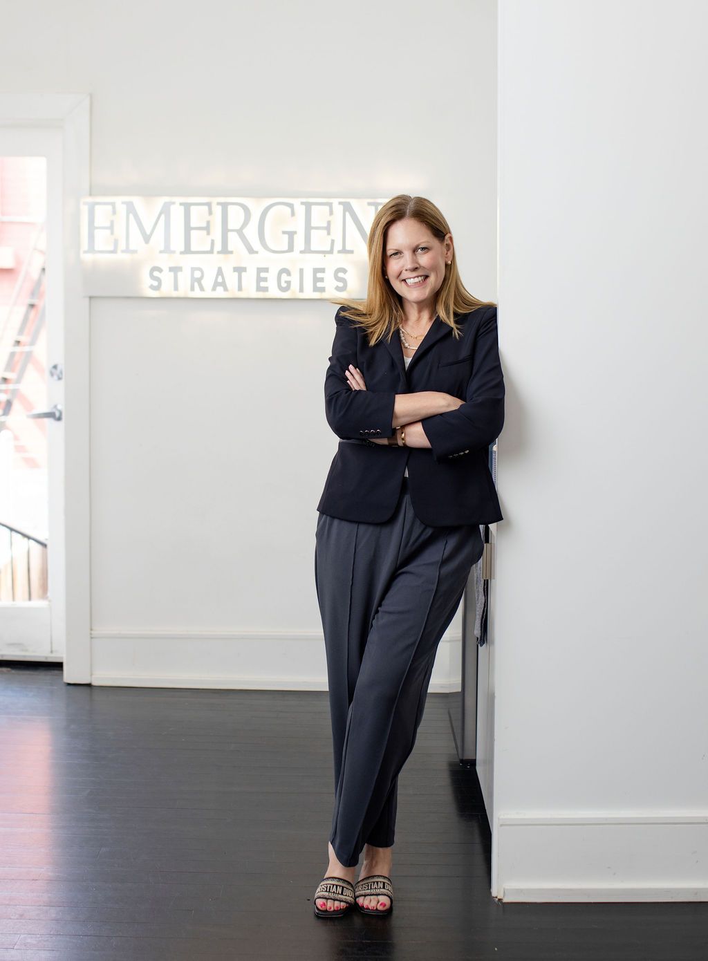 Professional woman in navy blazer poses confidently at Emergence Strategies office, arms crossed, smiling warmly.