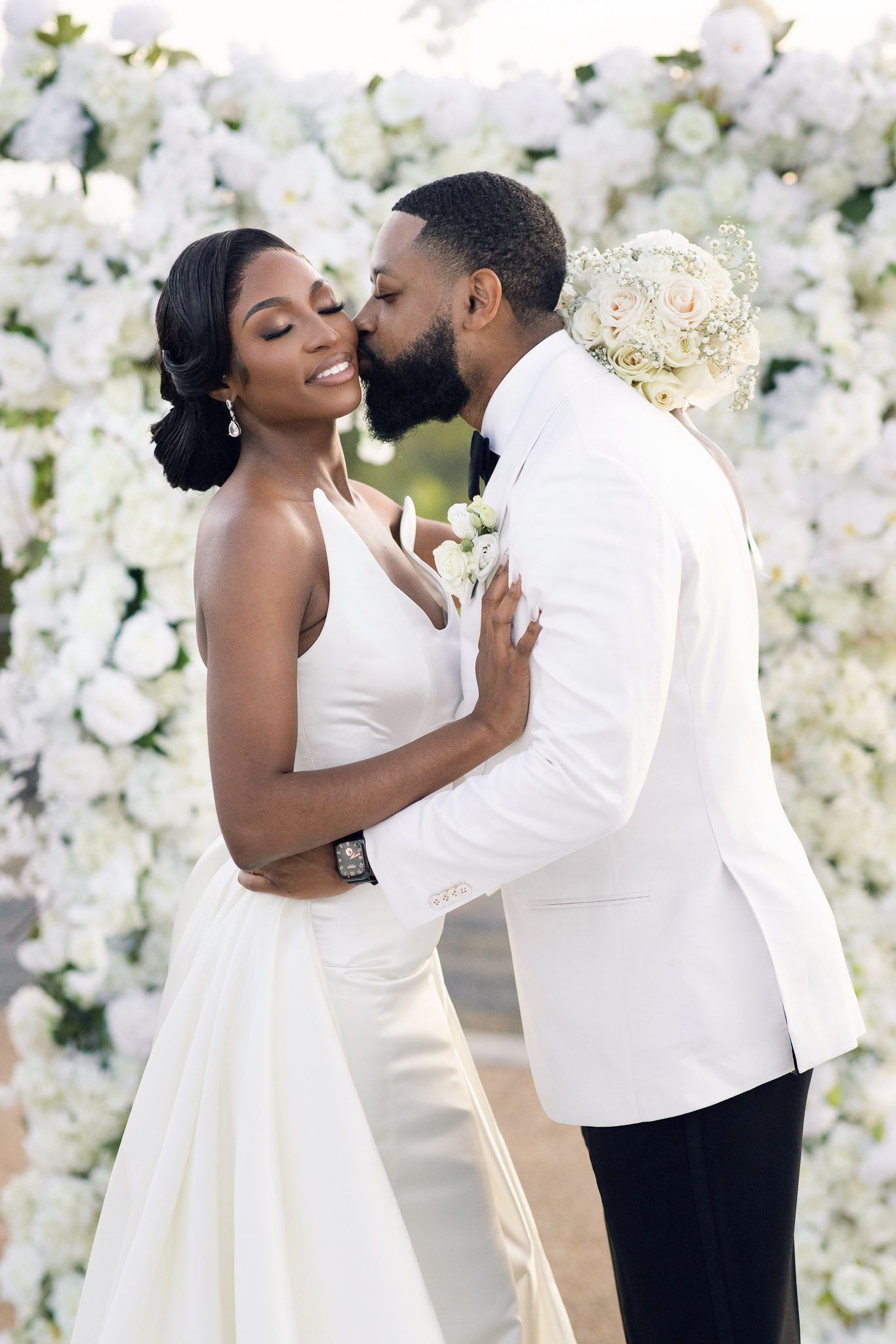 Joyful couple in white wedding attire share tender moment by floral wall.