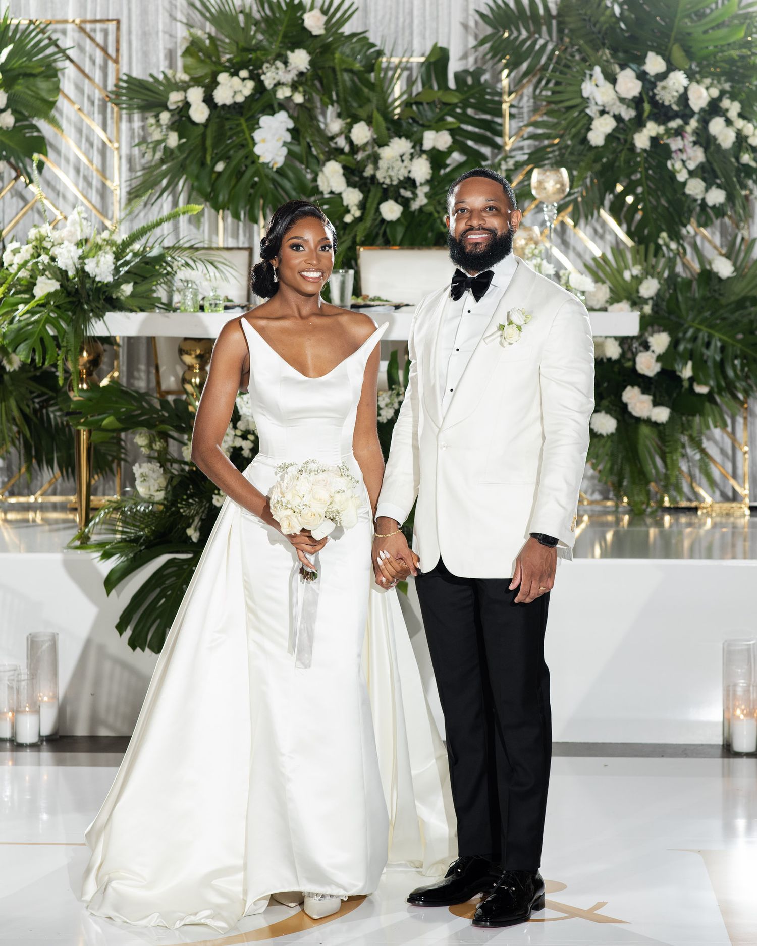 Elegant wedding ceremony setup with white floral arch, palm leaves and romantic decor in an indoor venue.