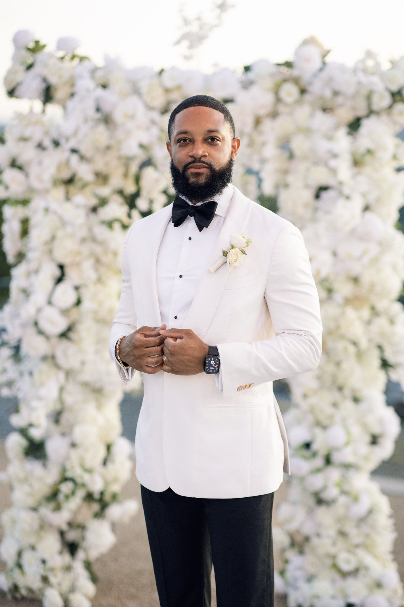 Elegant groom in white tuxedo jacket and black bow tie poses against floral wall backdrop.