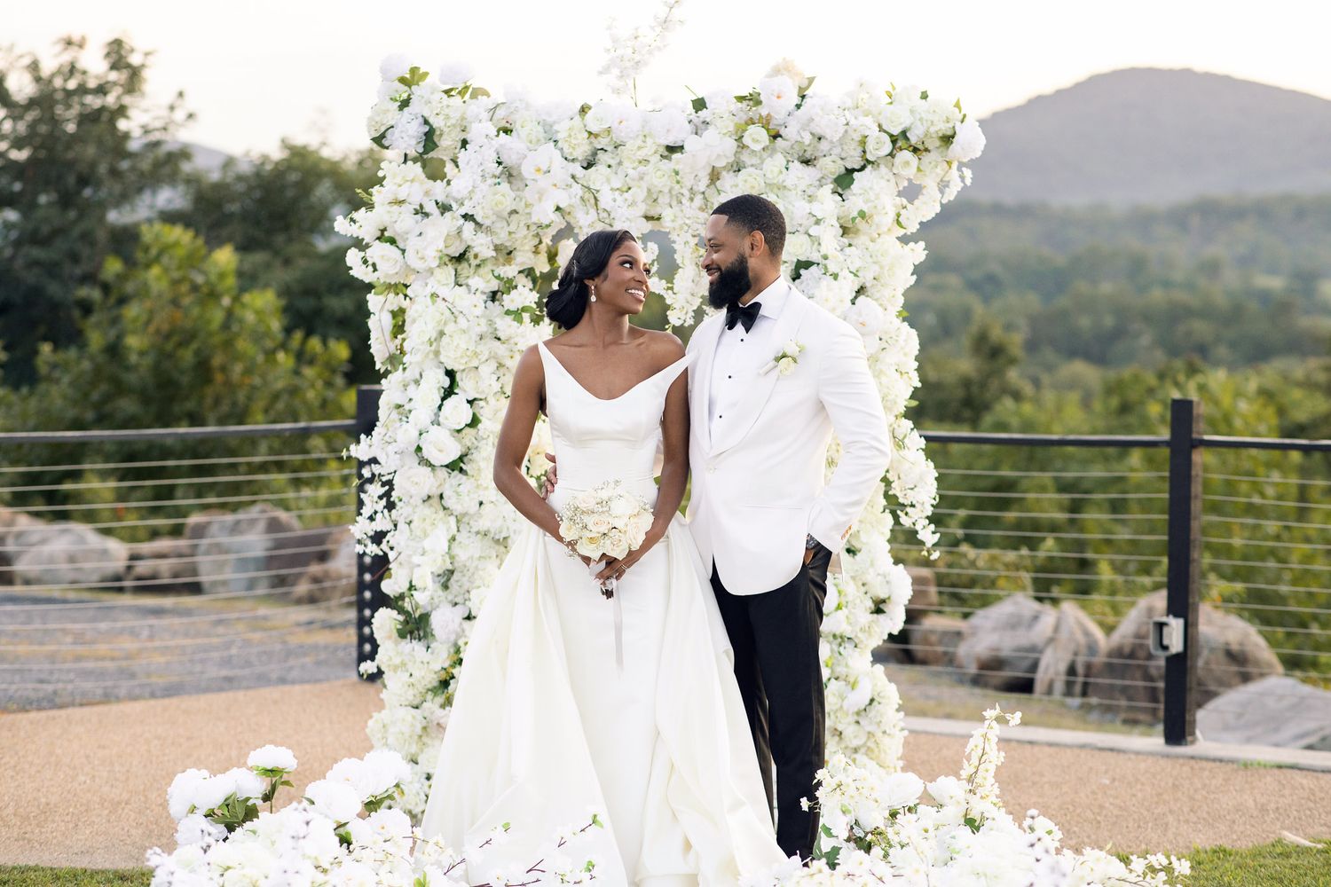 Newlyweds stand together under beautiful white floral arch with mountain view.