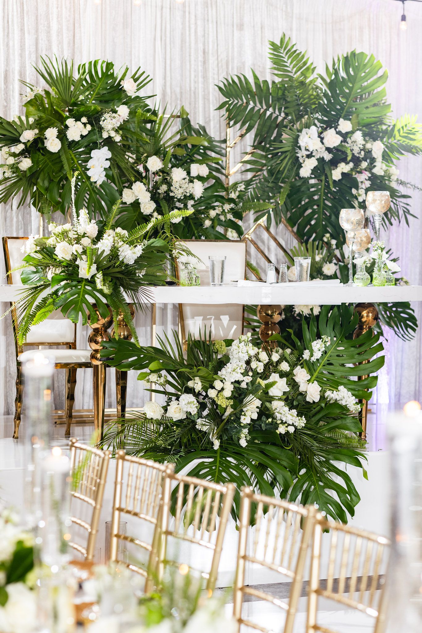 Tropical greenery and white floral arrangements line gold chairs at a wedding reception.