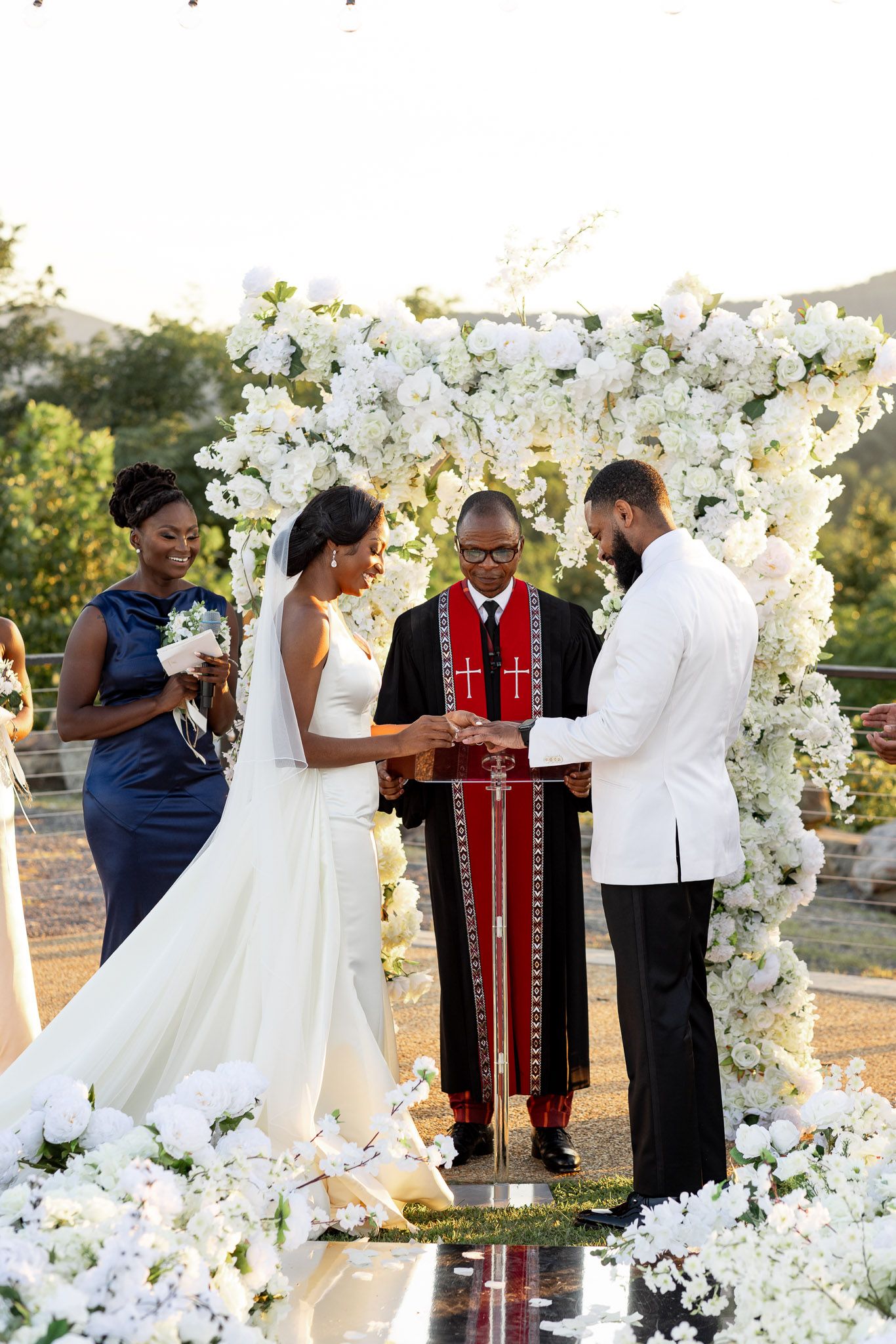 A wedding ceremony under a white floral arch with officiant presiding over the exchange of vows.