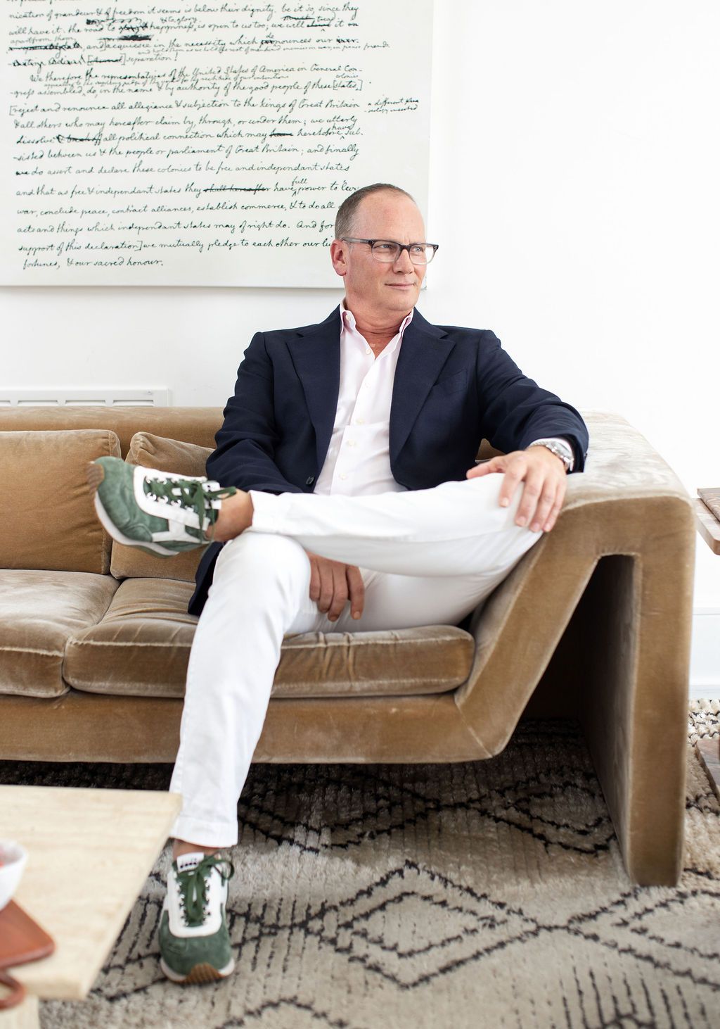 Man in navy blazer and white pants sits relaxed on a velvet sofa with green sneakers.