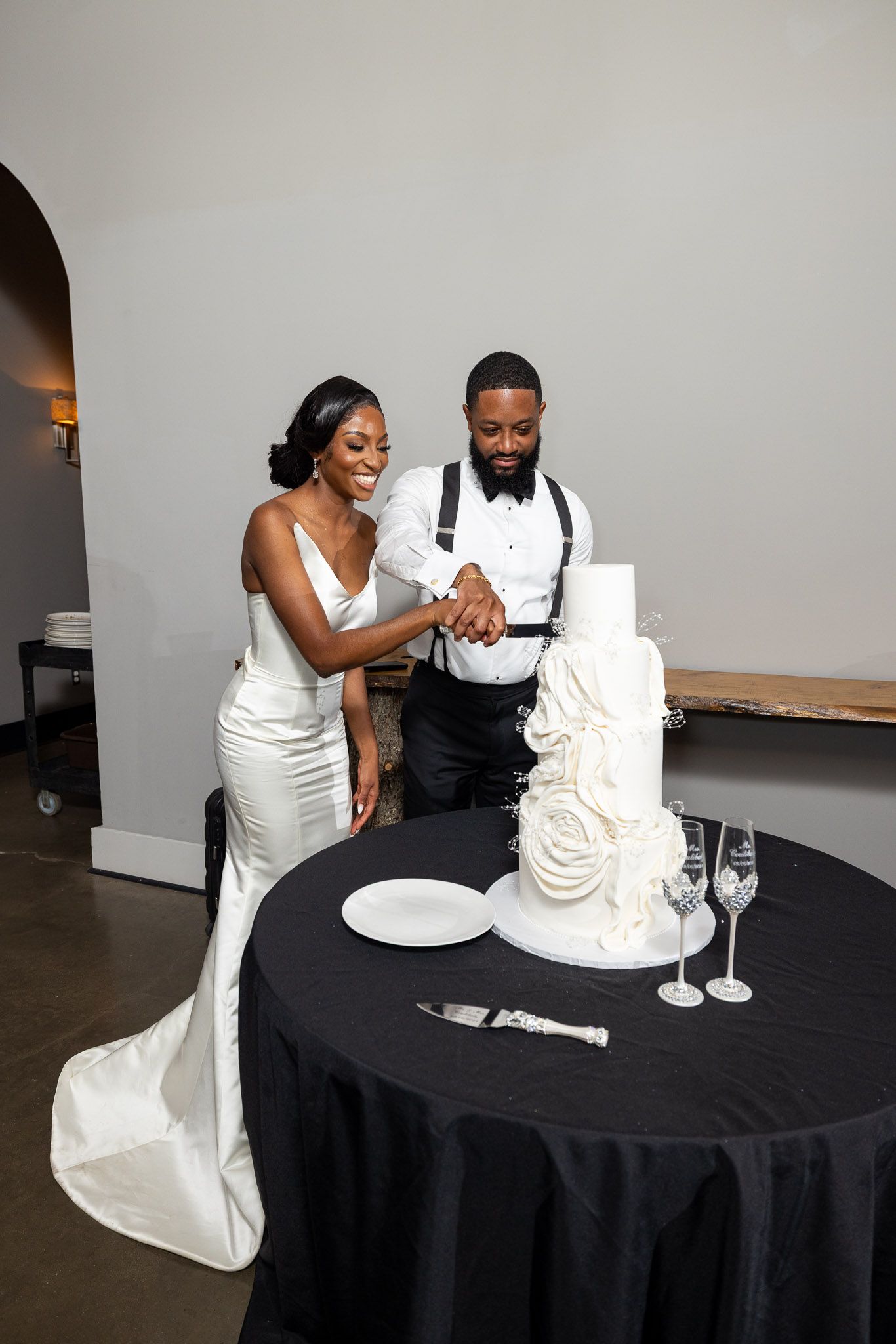Newlyweds share a joyful moment cutting their elegant white wedding cake adorned with floral details.