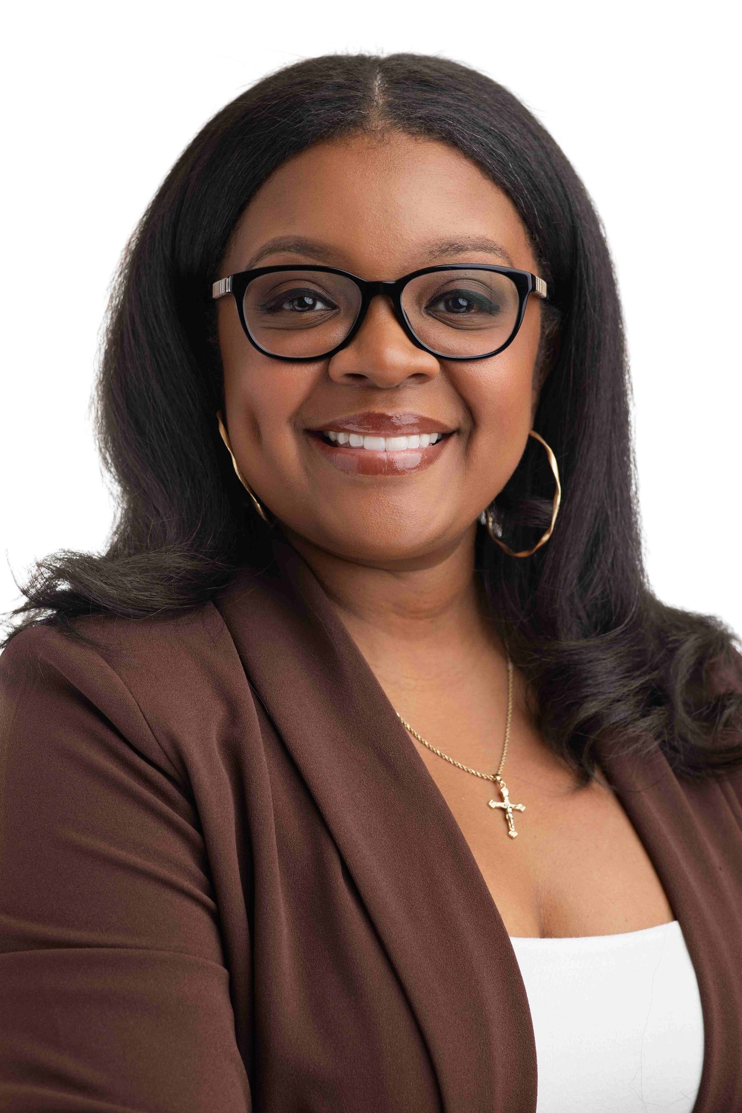 Professional headshot of a smiling woman wearing glasses, hoop earrings, cross necklace, and brown blazer on white background.