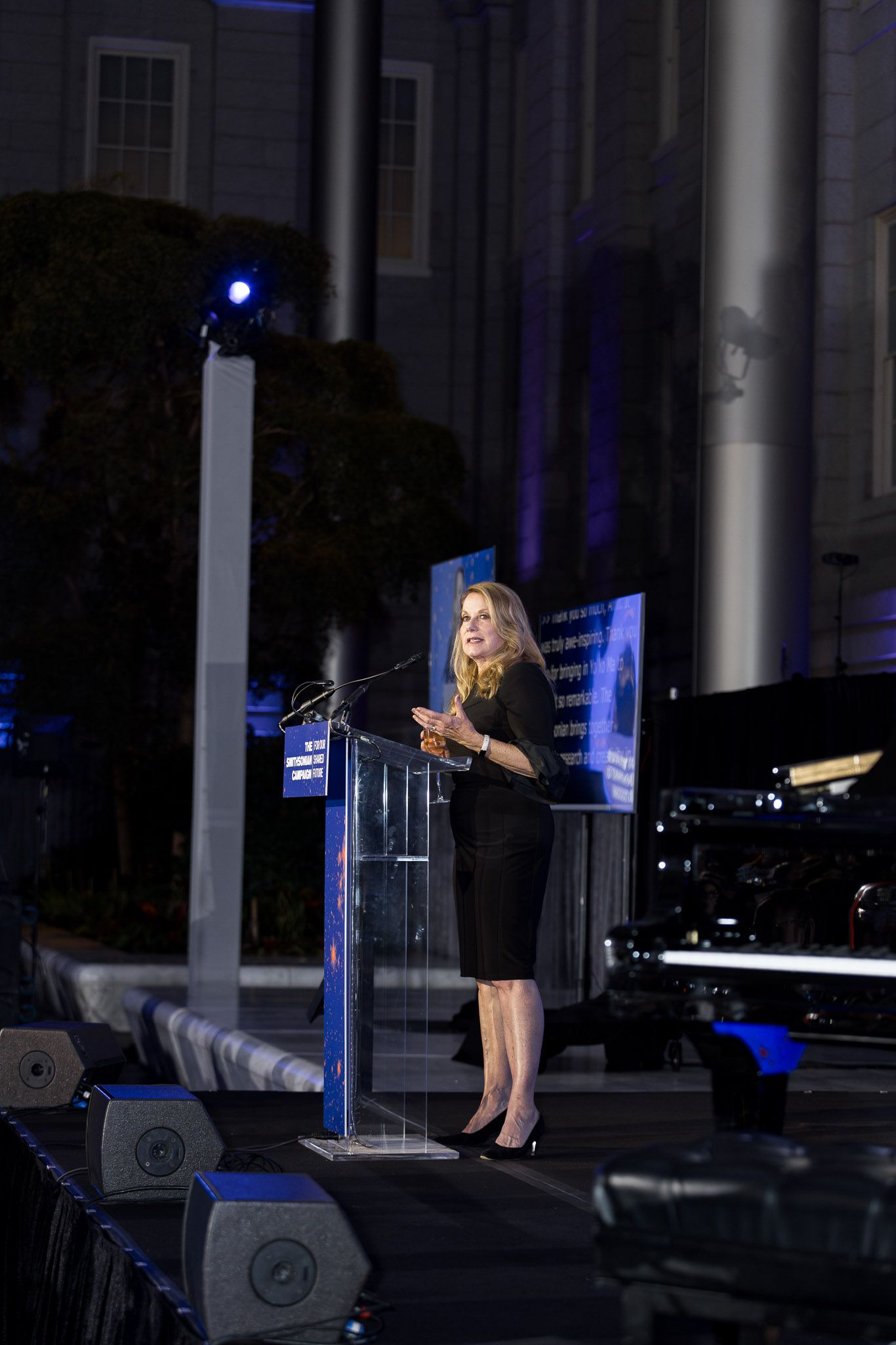 A person in a black dress stands at a podium giving a speech in a dimly lit venue with blue lighting.