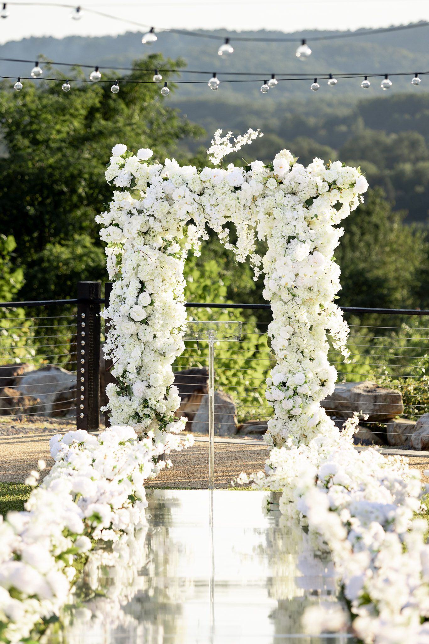 Wedding ceremony arch adorned with white flowers and hanging lights overlooking mountain scenery.