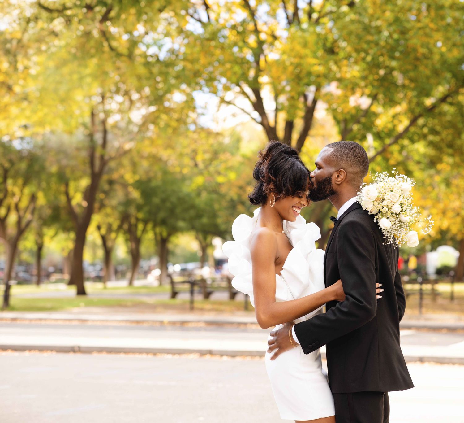 A Black bride and groom share a romantic moment outdoors, surrounded by autumn trees, as she holds a white bouquet.