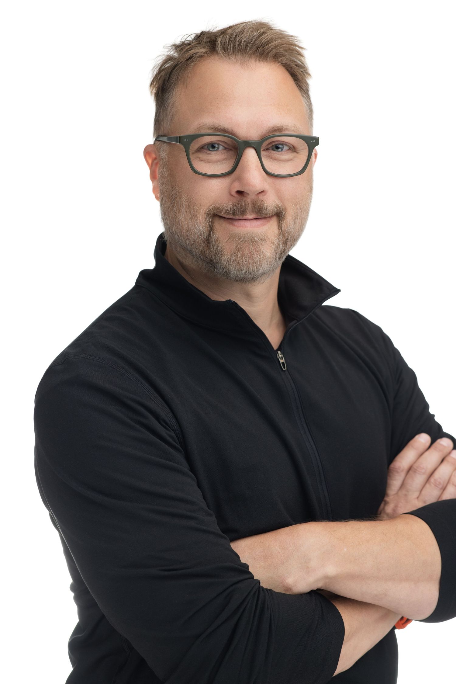 Middle-aged man with glasses and beard wearing black jacket, arms crossed, smiling confidently against white background.