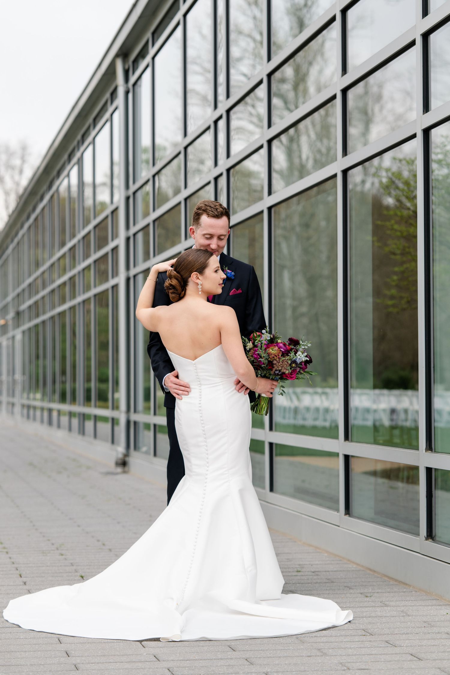 Elegant wedding photos of couple by modern glass building with bride's dress trailing behind.