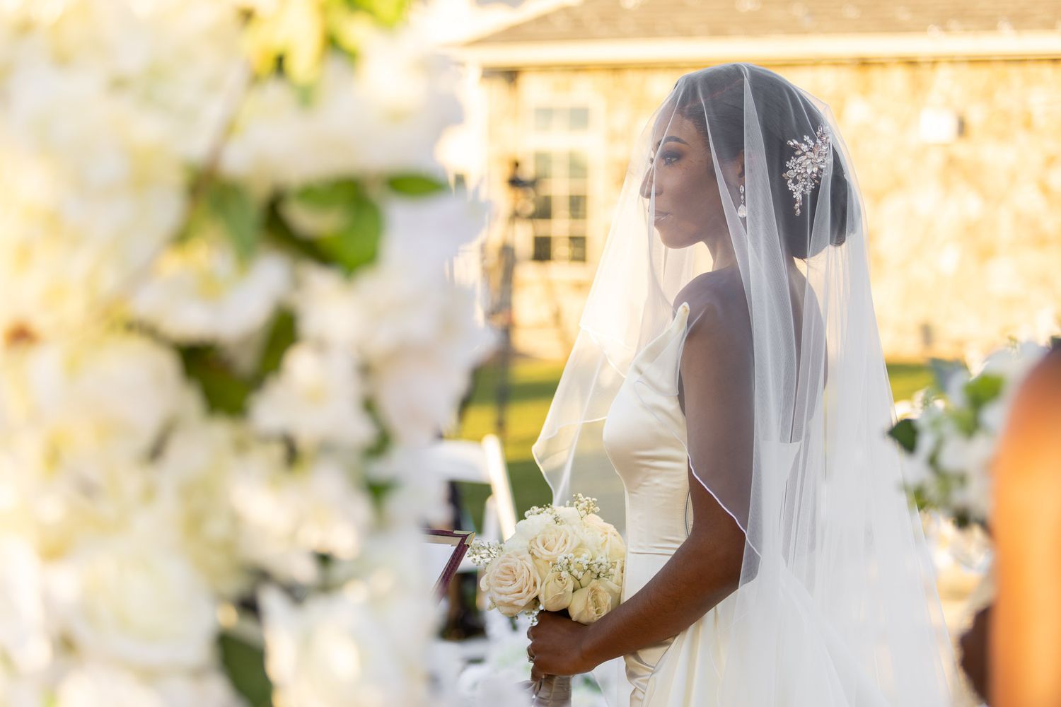 Bride in elegant white gown and veil holding bouquet during golden hour ceremony.