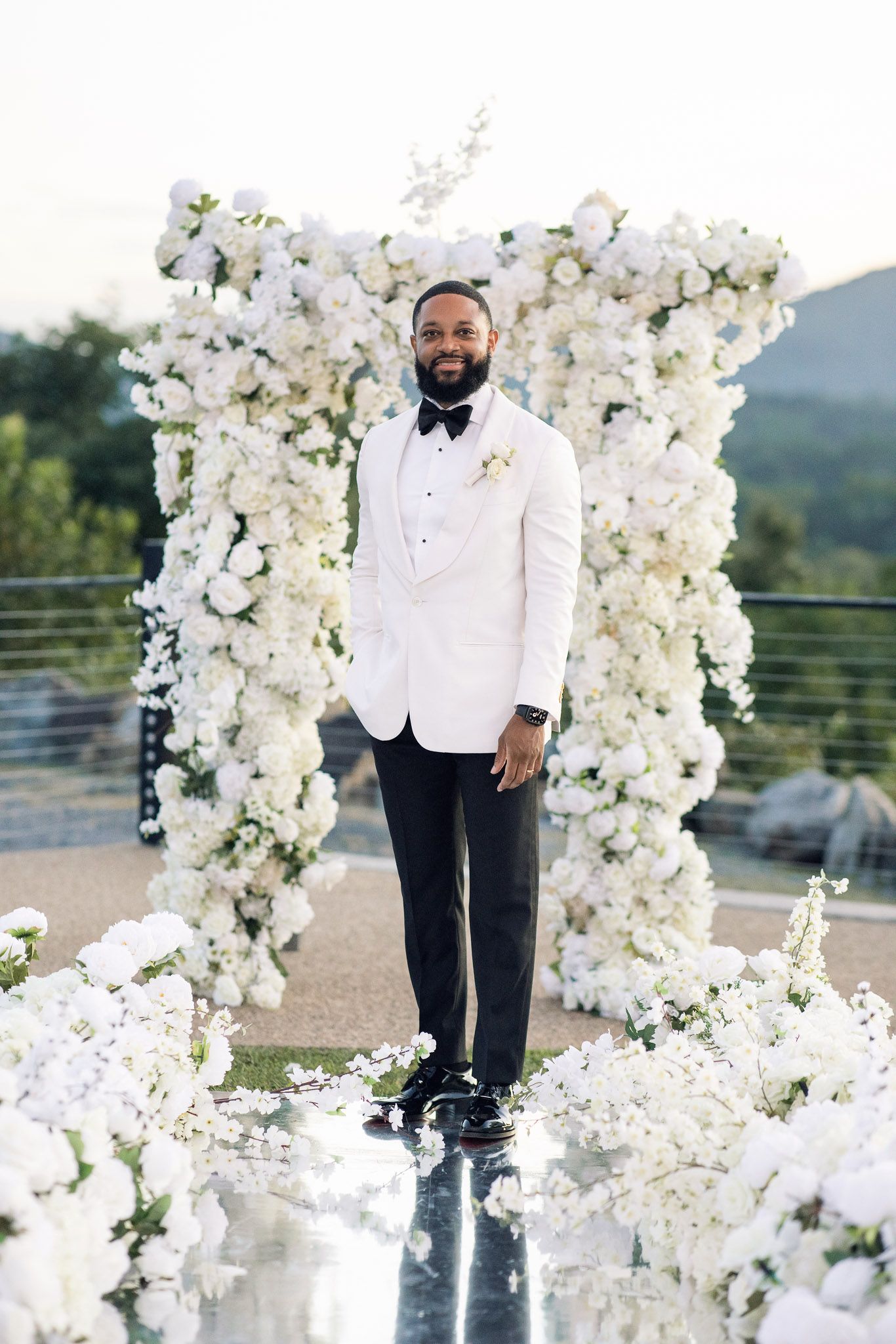 Groom in white tuxedo jacket and black bow tie poses by luxurious floral arch installation.