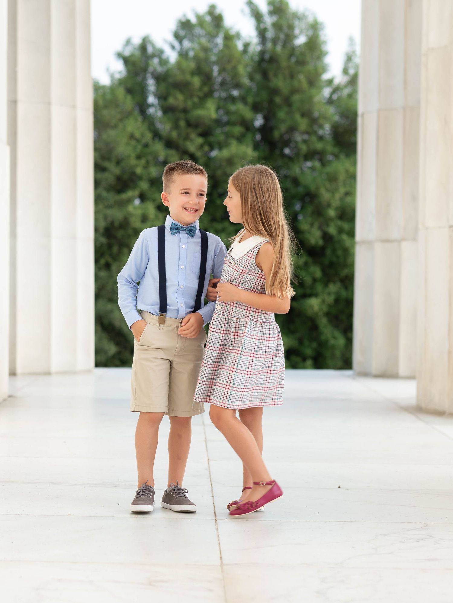 Two dressed-up children in suspenders and plaid dress pose between white columns outdoors.