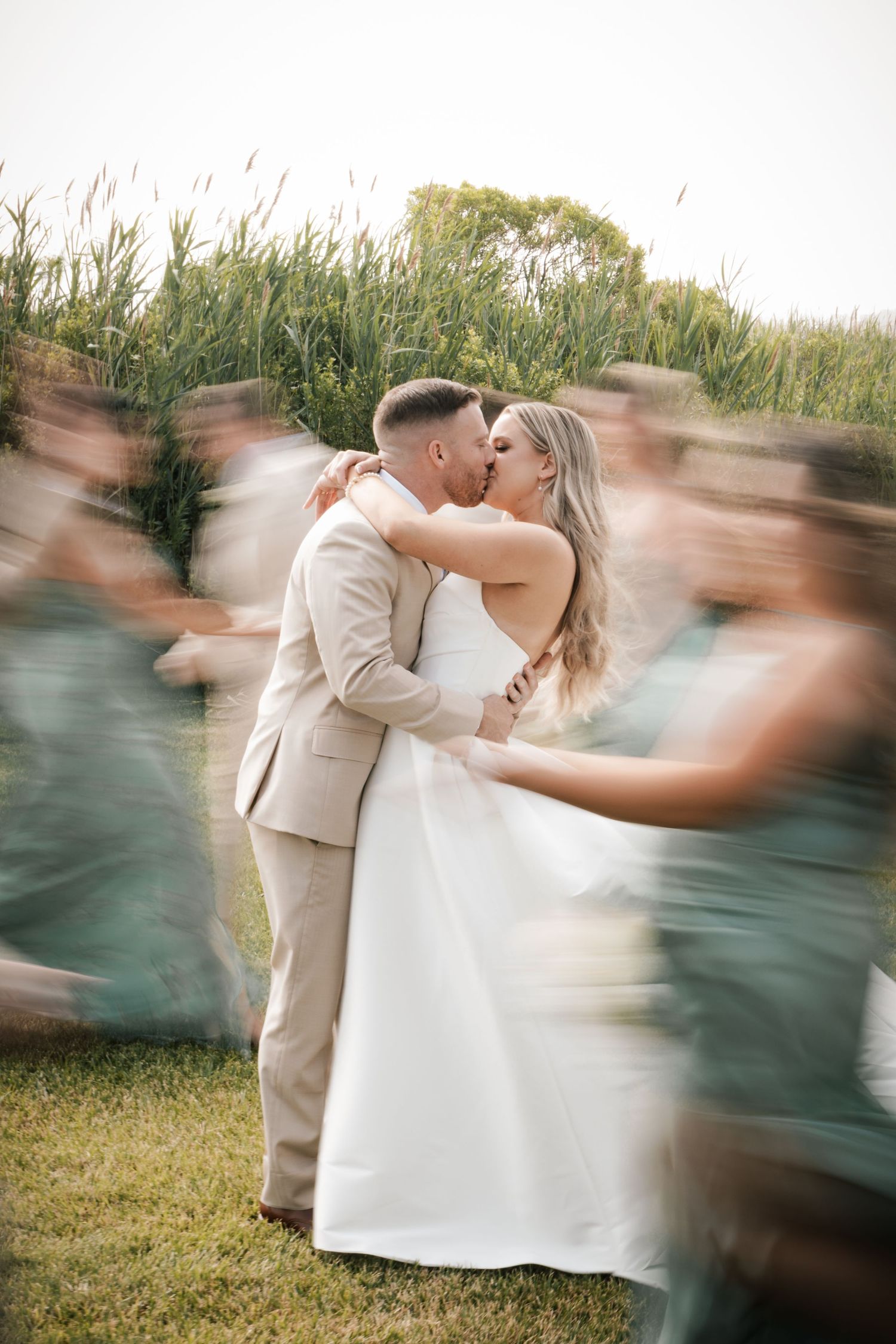 A bride and groom share a passionate kiss surrounded by blurred motion of wedding party on a lawn in New Jersey.