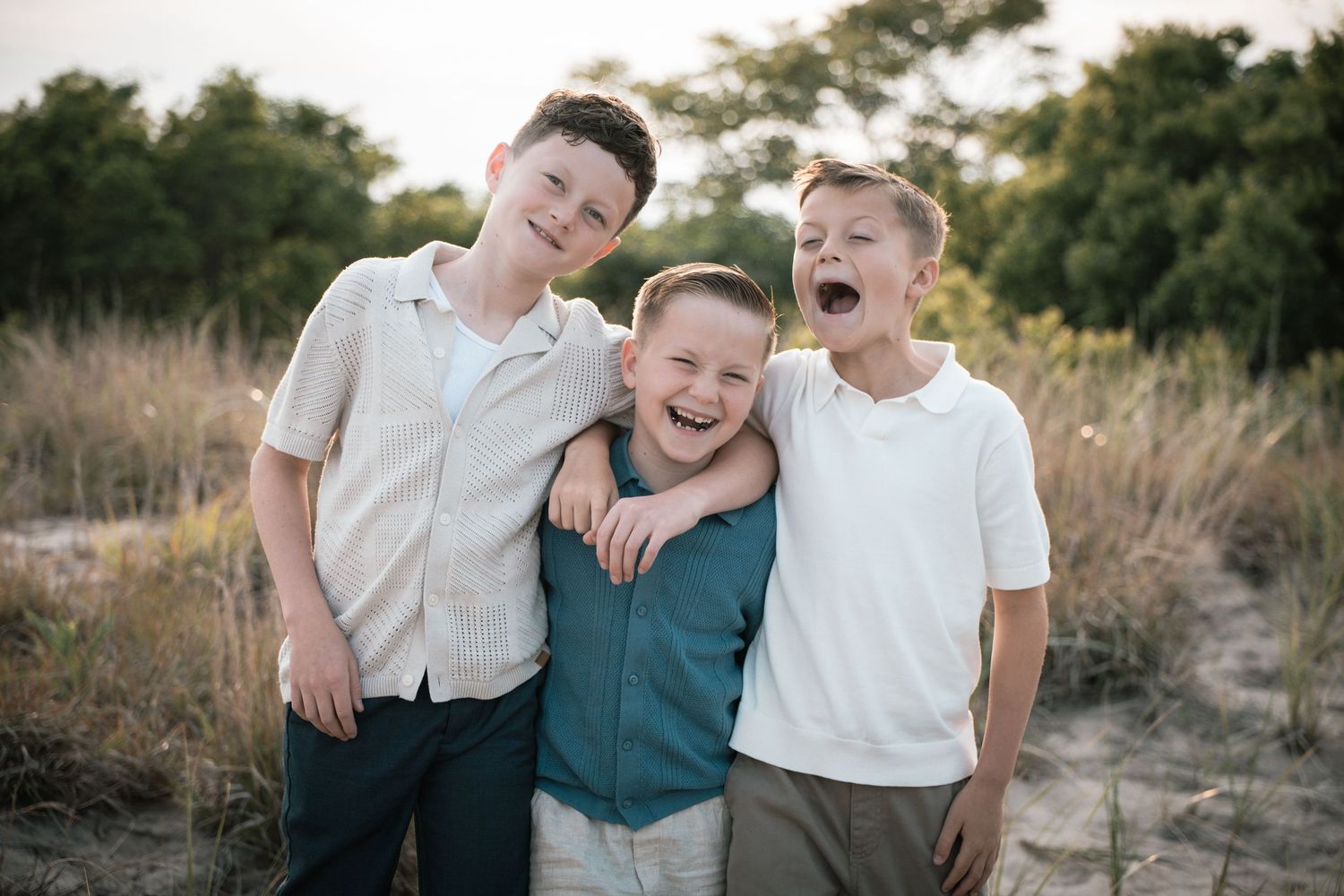 Three young boys laugh and hug each other outdoors in a sun-drenched natural setting.