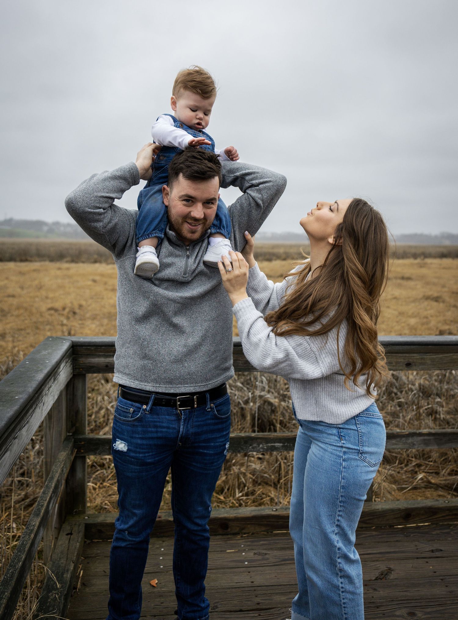 Young couple on wooden bridge lifting baby boy overhead against cloudy autumn field backdrop.