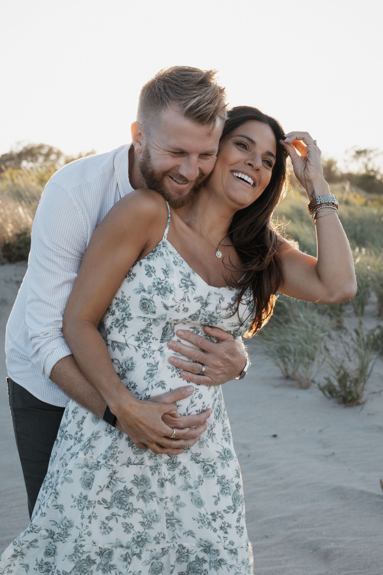Smiling couple embracing on sandy beach dunes at golden hour, woman in green floral dress.