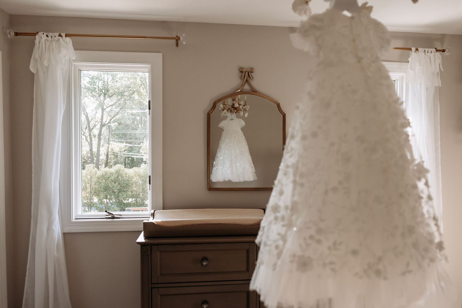 White lace christening gown hanging in neutral-toned nursery room with wooden dresser and sheer curtains.