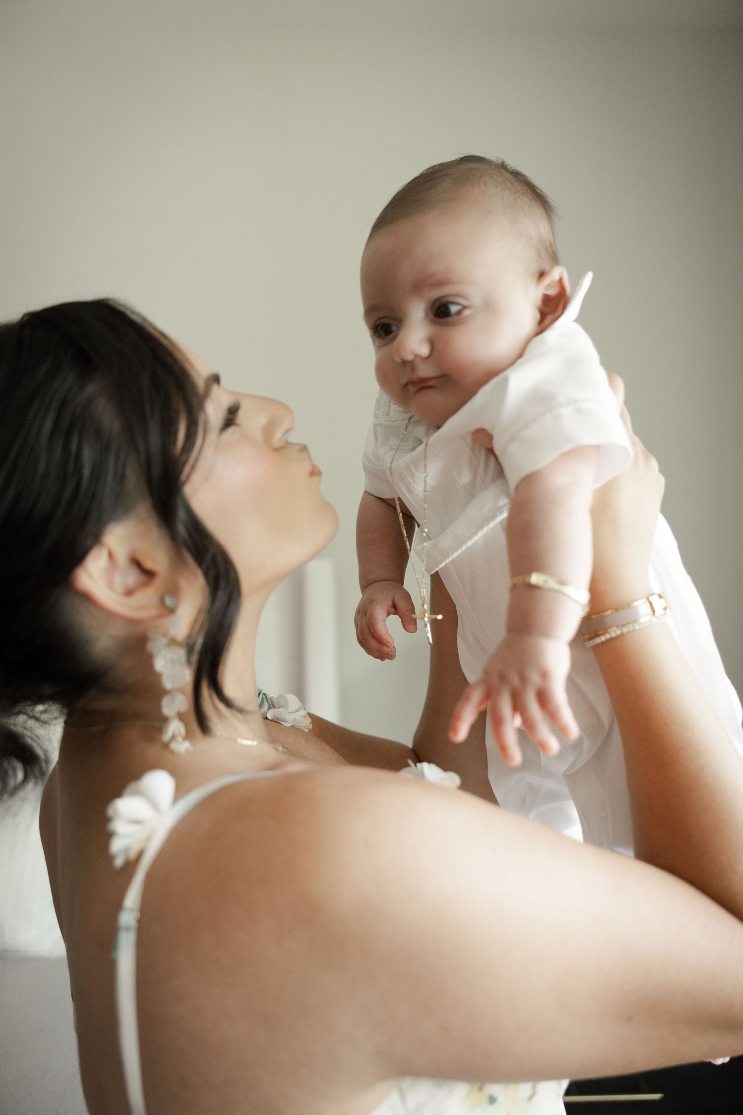 Mother lifting smiling baby in white christening outfit with joy.