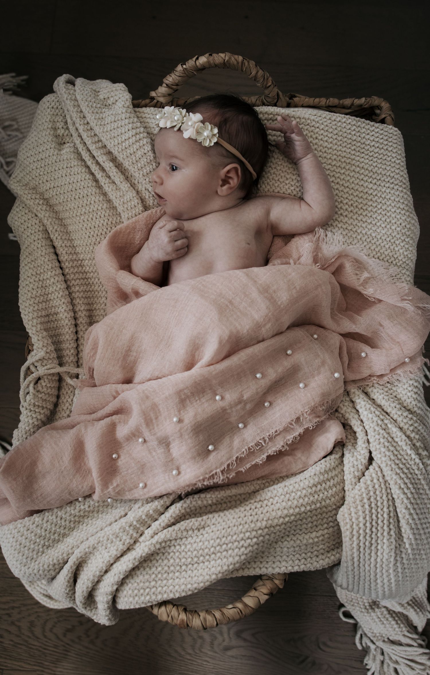 Sleeping newborn baby in white flower headband wrapped in pink pearl muslin blanket in wicker basket.