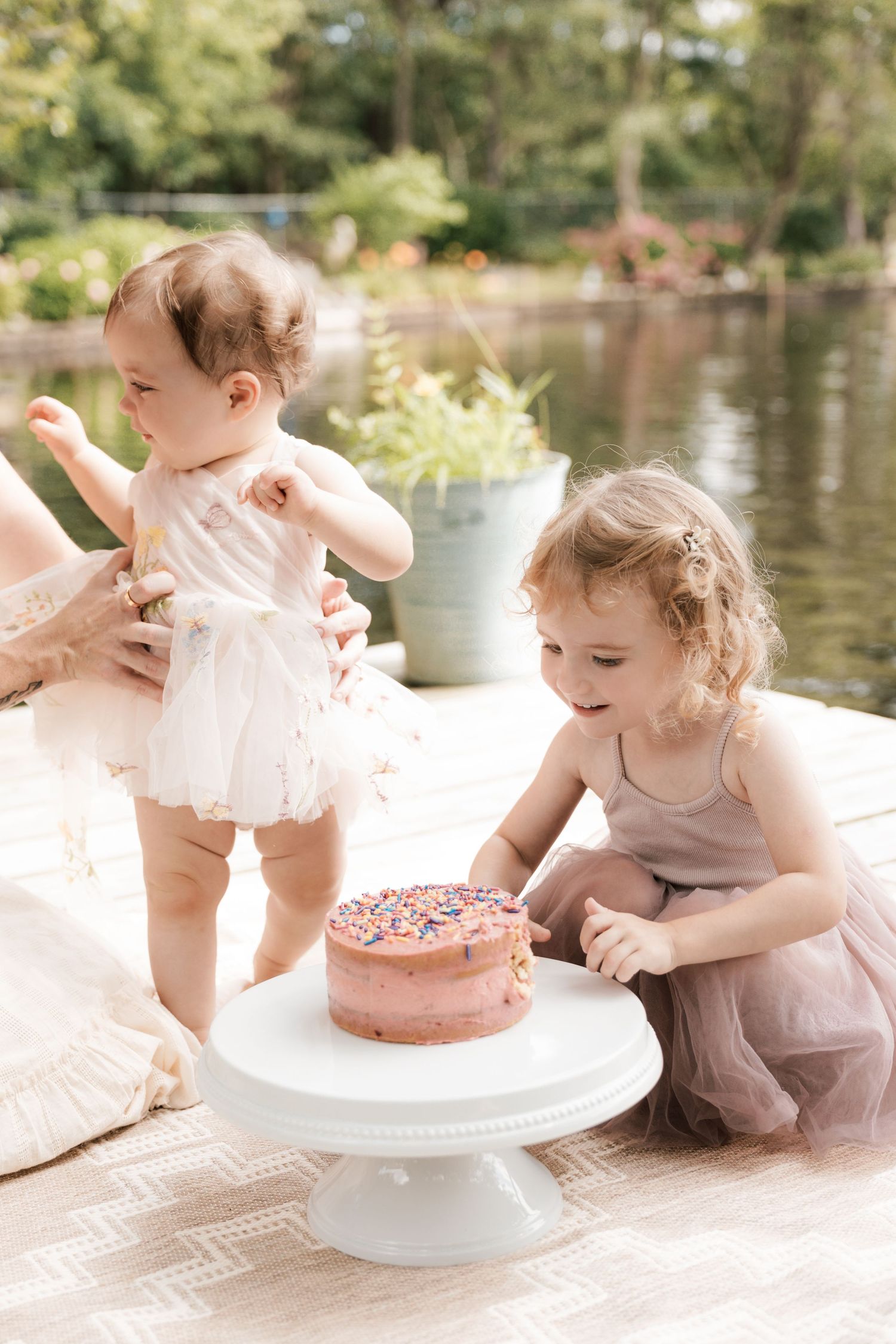 Two little sisters in pink dresses look at sprinkle birthday cake on dock.