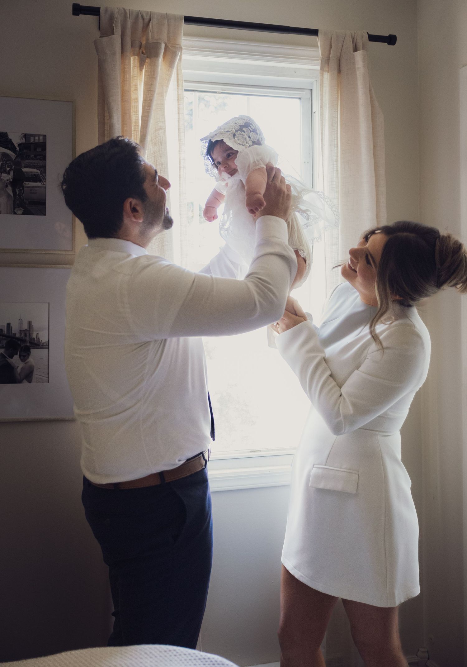 Parents joyfully lifting their baby in a white christening gown near a bright window in a cozy room.