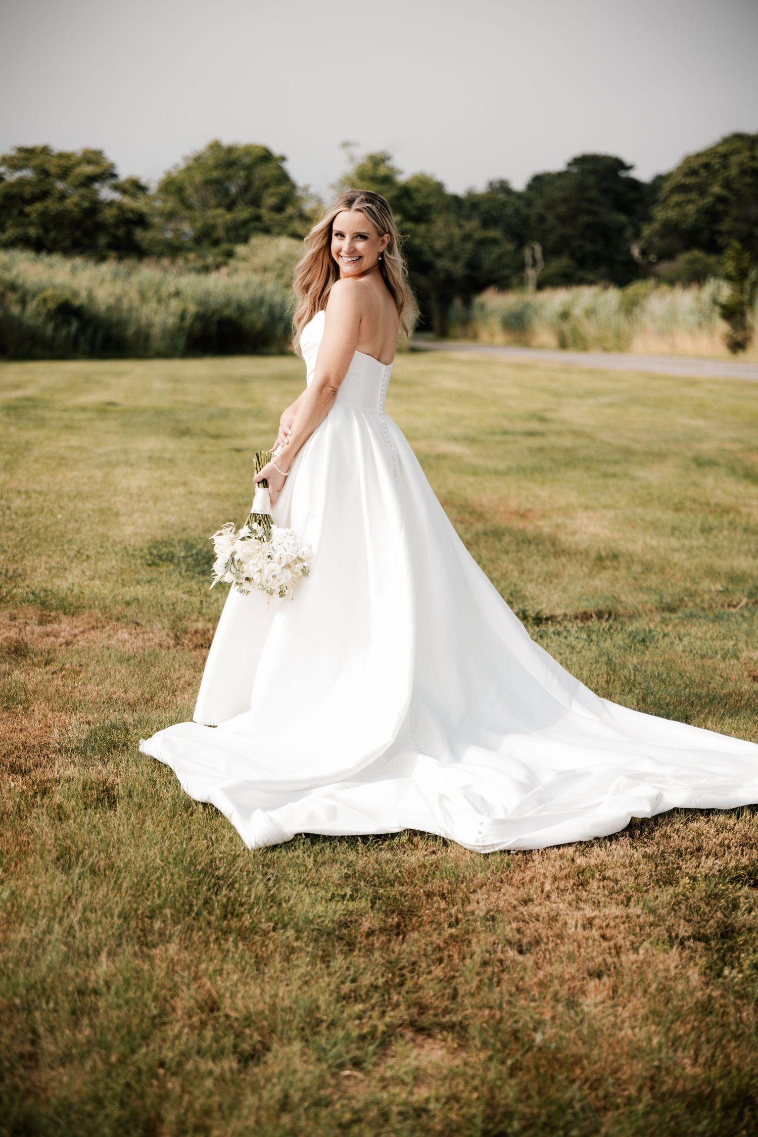 An elegant bride in a flowing white strapless gown with a long train smiles in a green meadow in New Jersey.