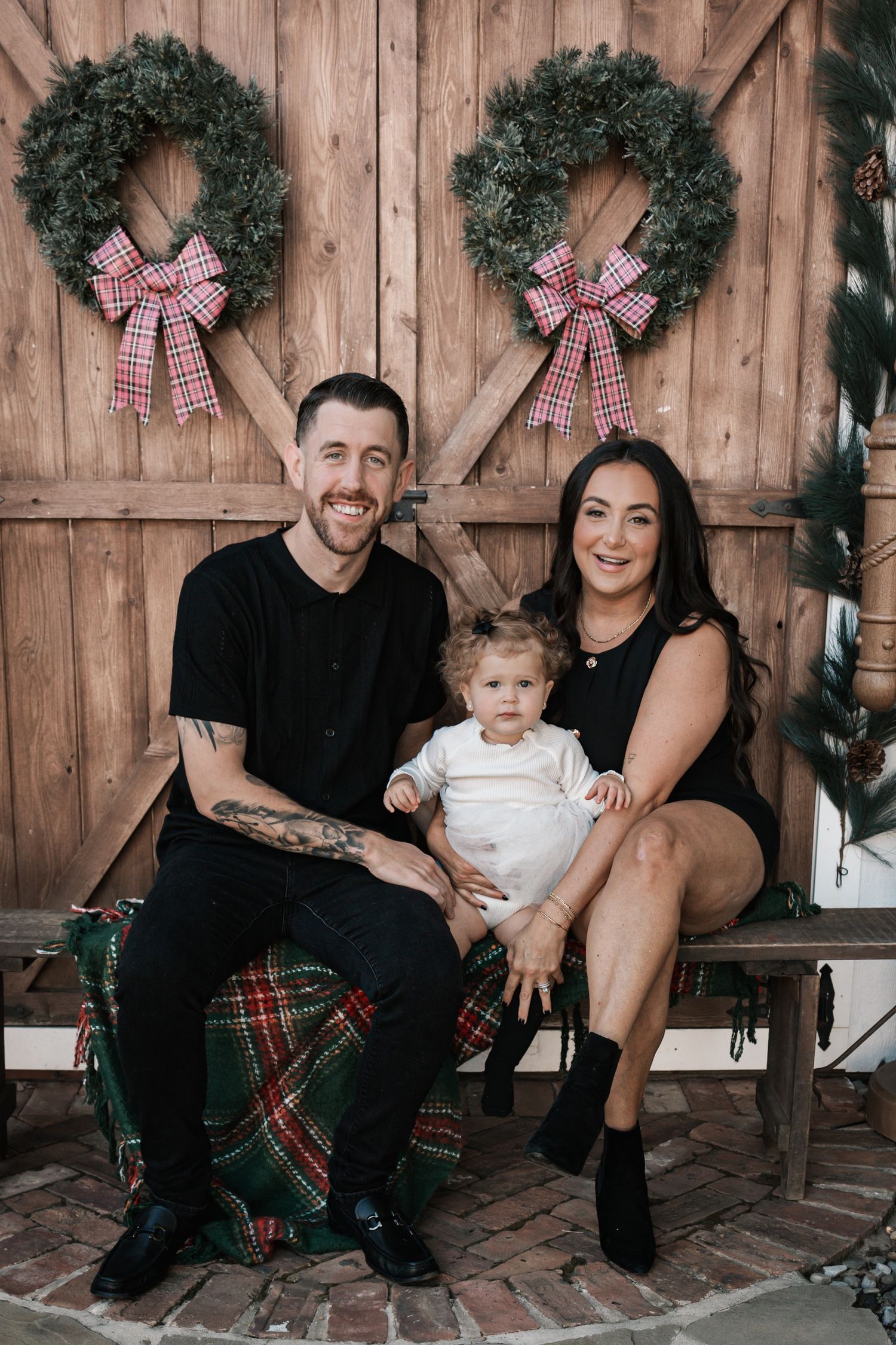 Happy family of three posing on bench before rustic barn doors decorated with Christmas wreaths and red bows.