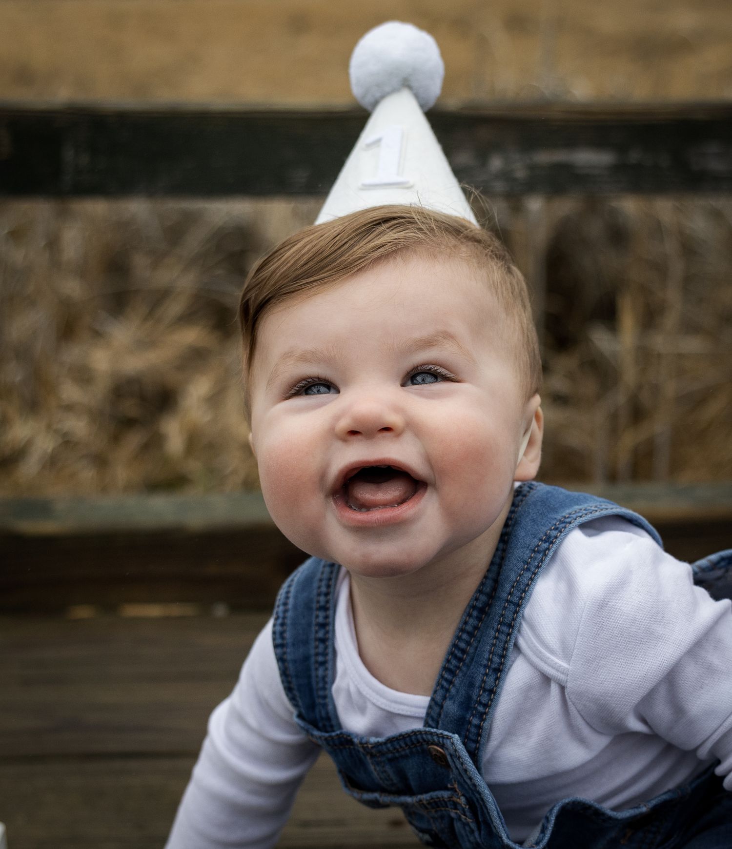Laughing baby boy wearing white party hat and denim overalls crawling on rustic wooden surface.