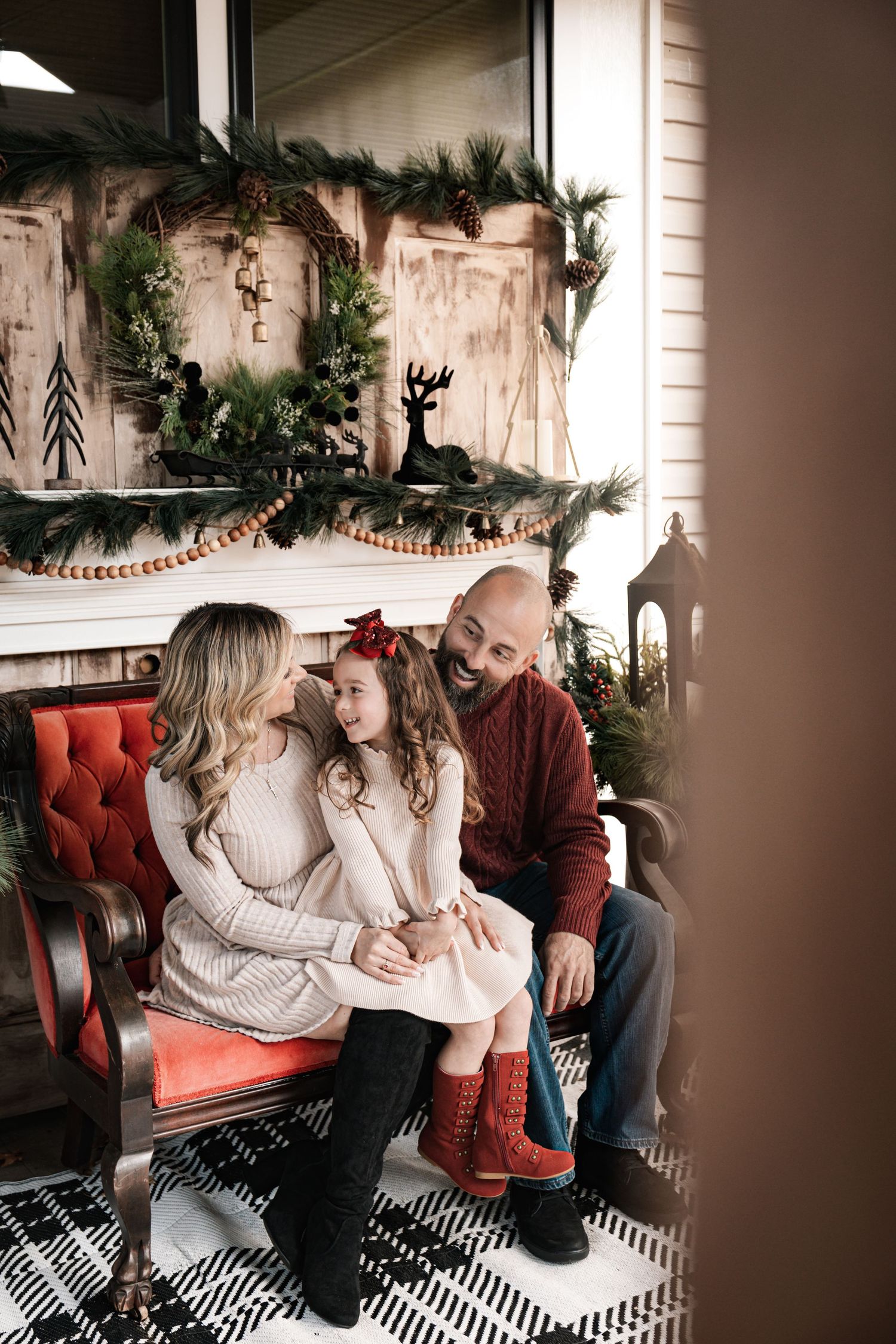 A family of three sits together on a red velvet bench beside a Christmas fireplace mantel.