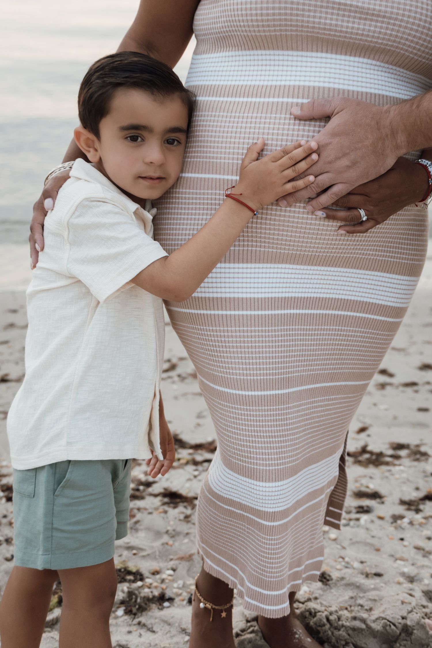 Young boy hugging pregnant mothers striped belly on sandy beach during family maternity session.