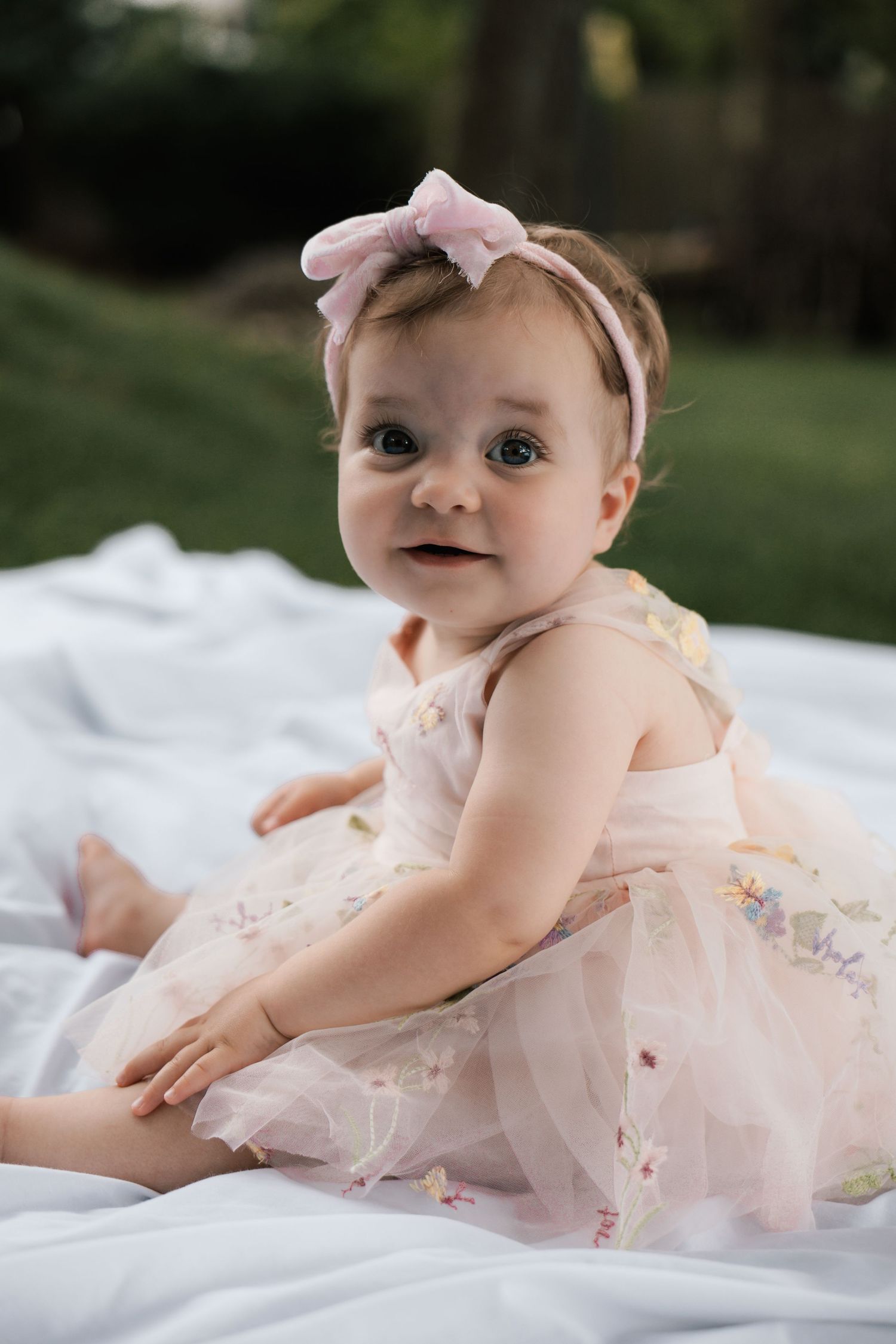 Cute baby girl in floral pink tutu dress with pink bow headband sitting on white blanket outdoors.