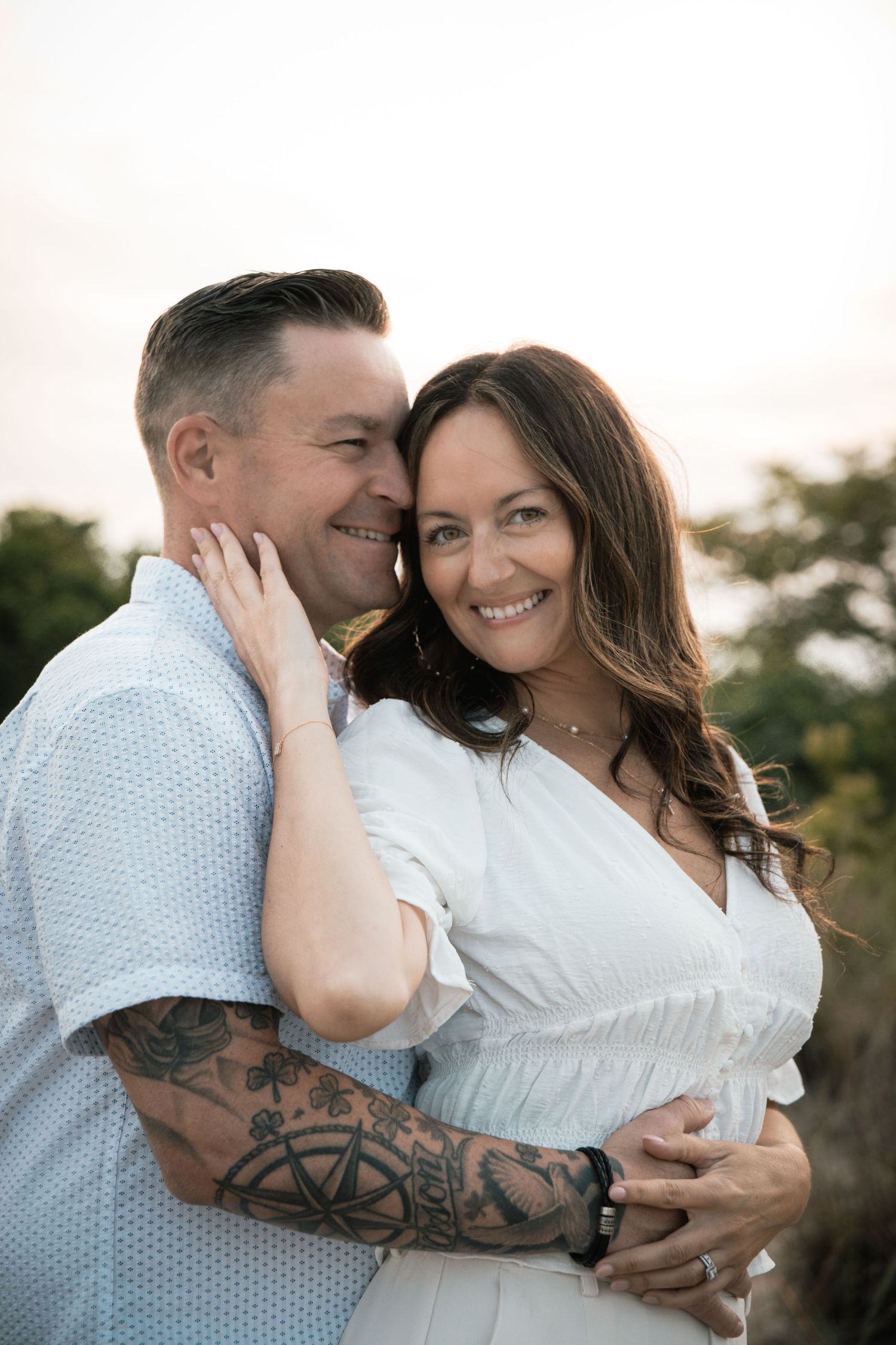 A smiling couple embraces outdoors at sunset, the man with tattooed arms hugging the woman.
