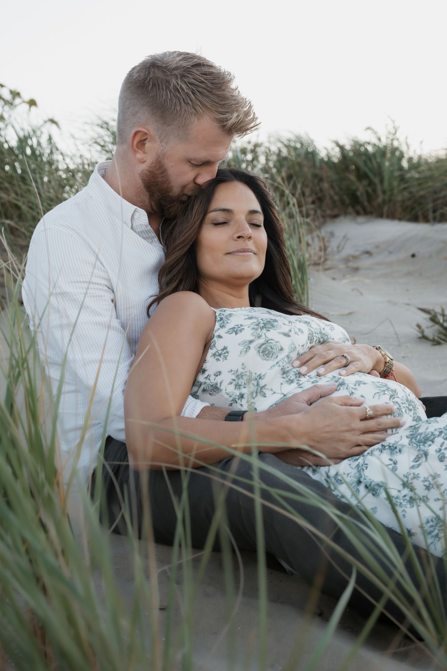 Couple sitting peacefully in beach sand dunes, man embracing pregnant woman in floral dress.