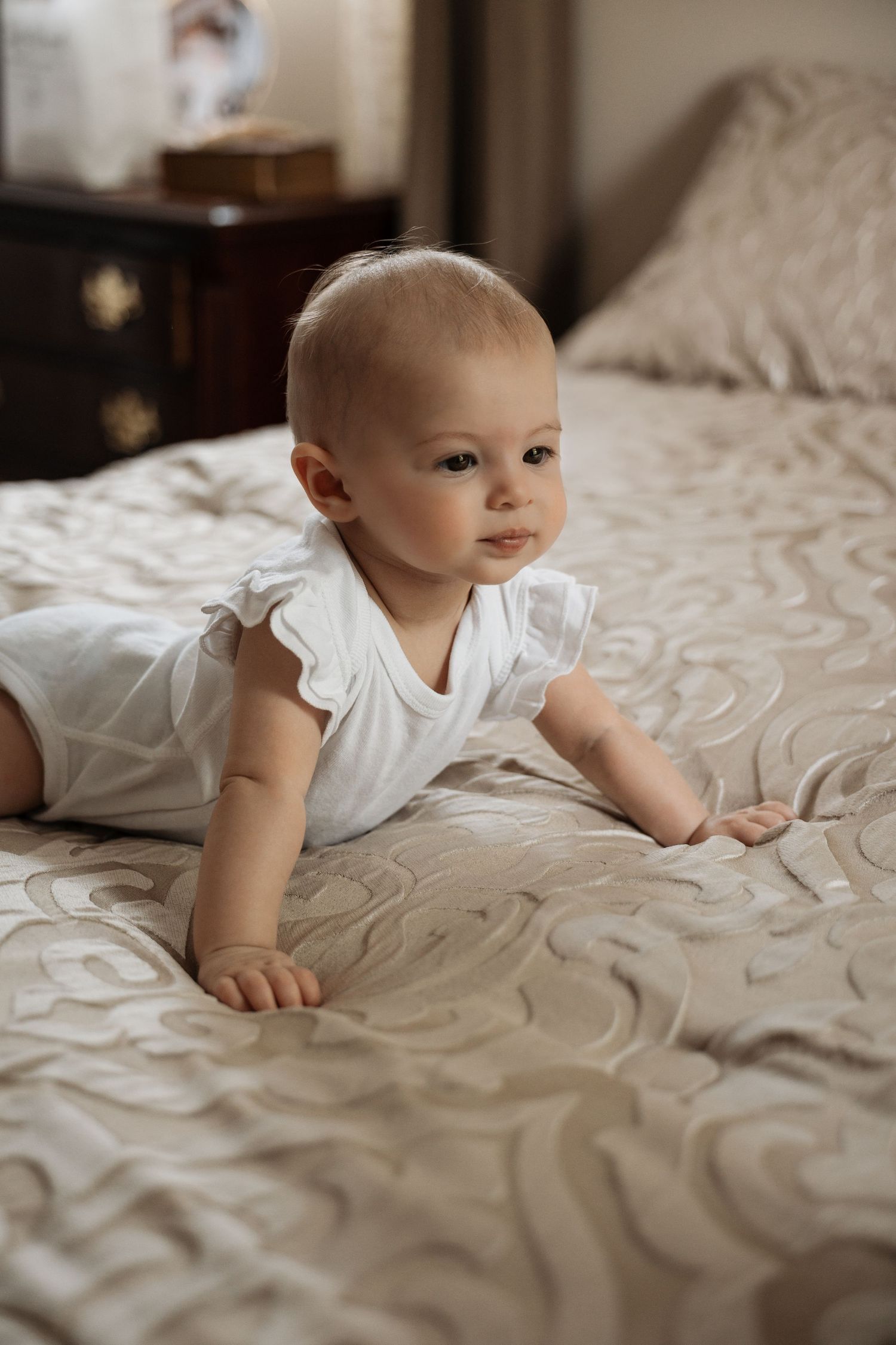 Cute baby in white ruffle onesie crawling on a beige patterned bedspread indoors.