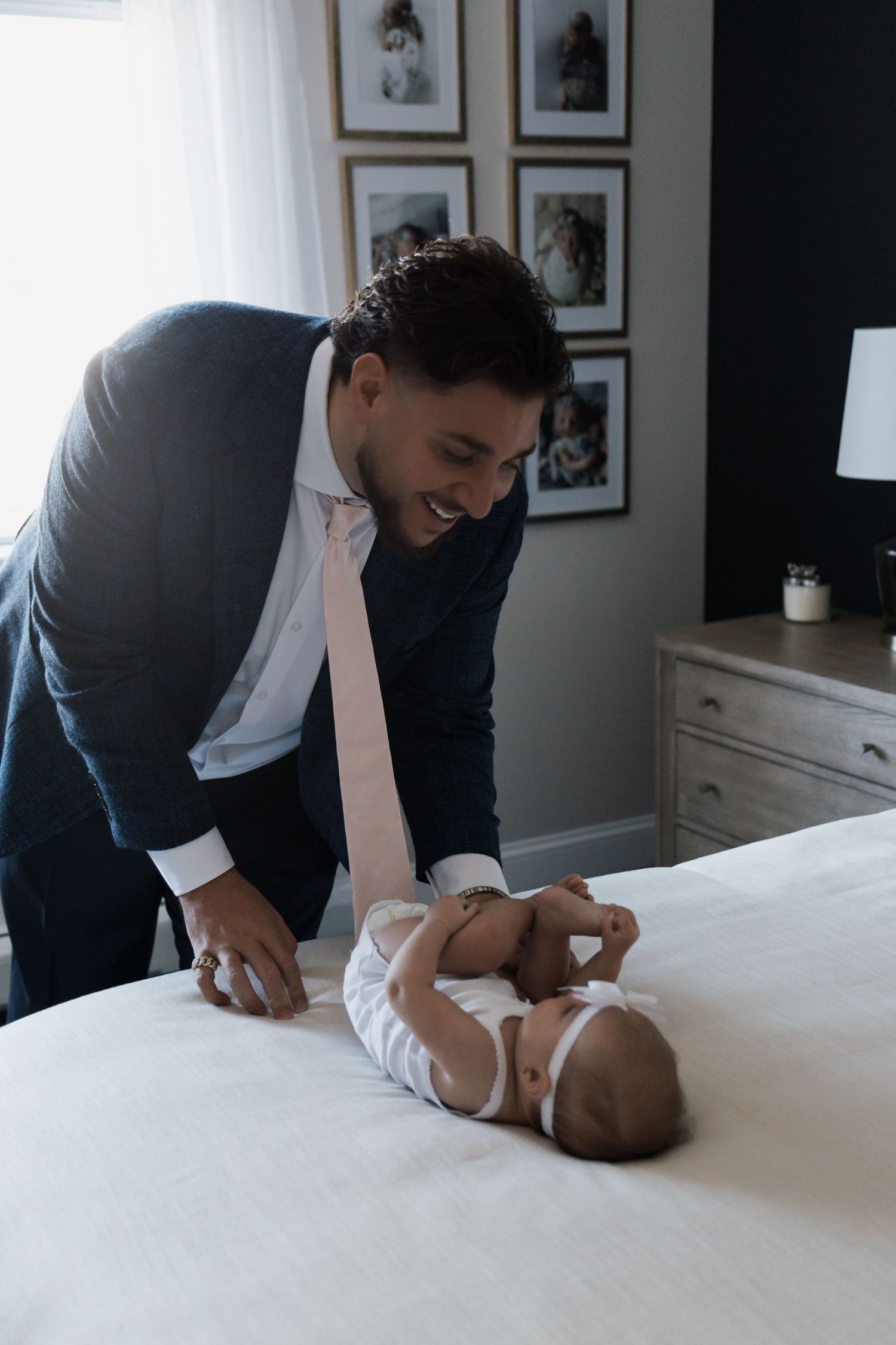 Smiling father in navy suit and pink tie leaning over laughing baby lying on white bed.