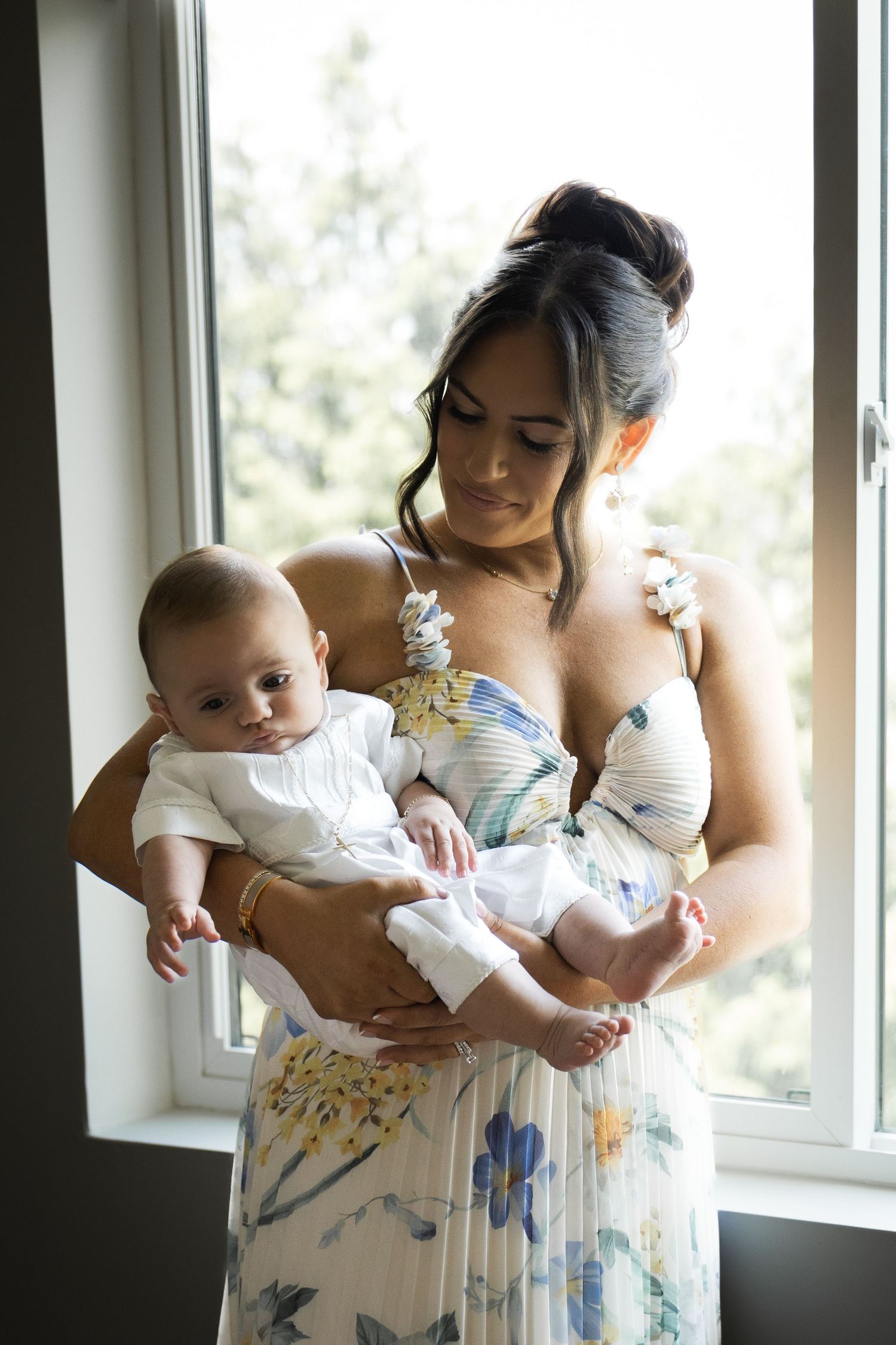 Elegant mother in floral dress holding baby in white christening outfit by window in Staten Island.