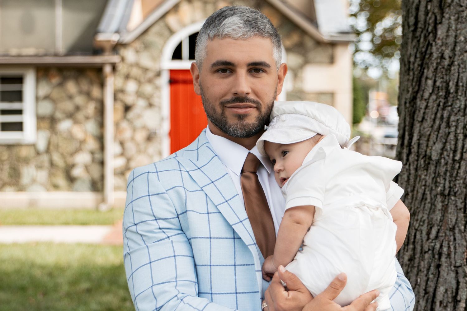 Father in light blue plaid suit holding baby in white christening outfit by a tree.