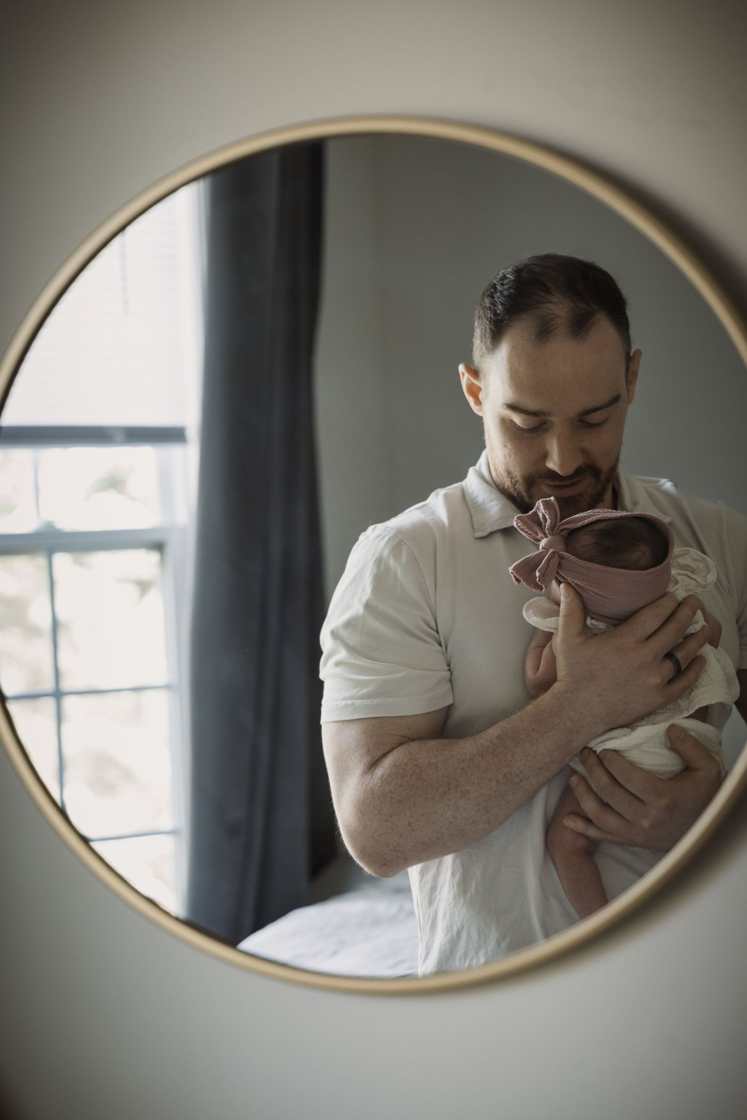 Father holding newborn baby with pink bow reflected in round gold-framed mirror near window.