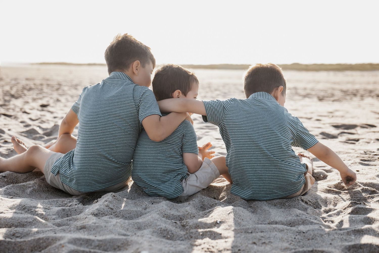 Three young boys in matching outfits sit together on a sandy beach, viewed from behind at sunset in New Jersey.