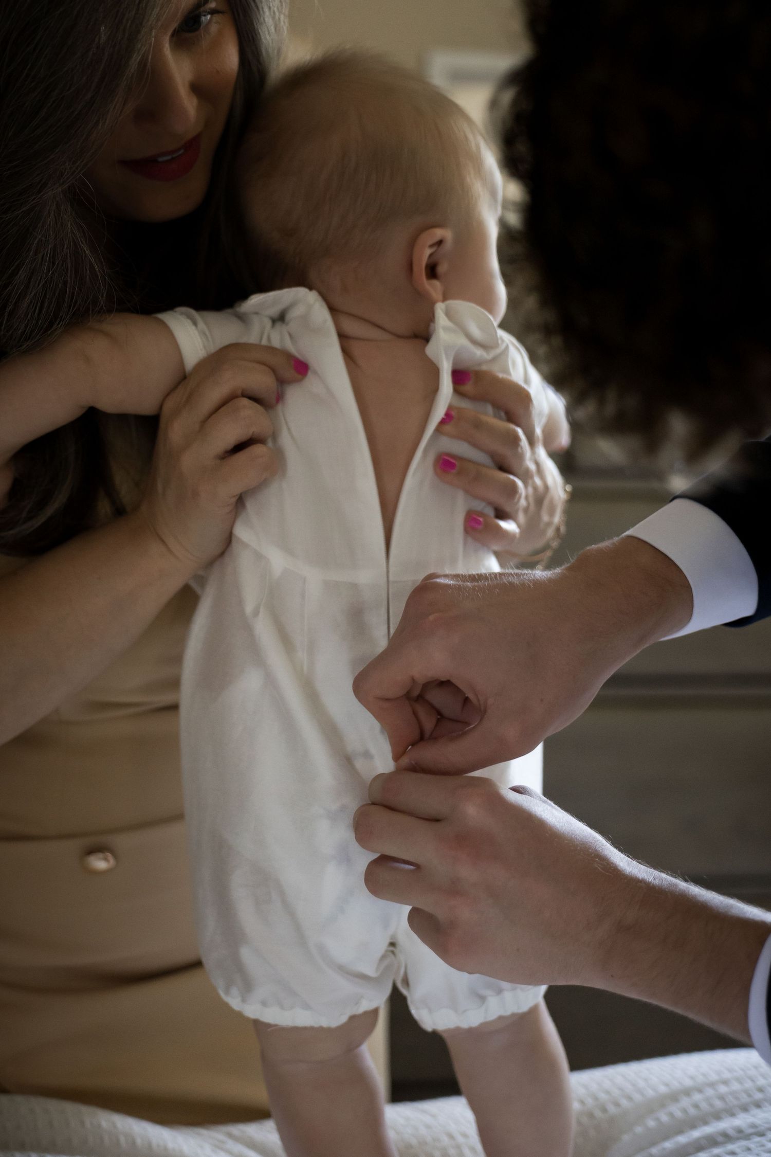 Adults carefully dressing a newborn baby in a white christening outfit on a bed.