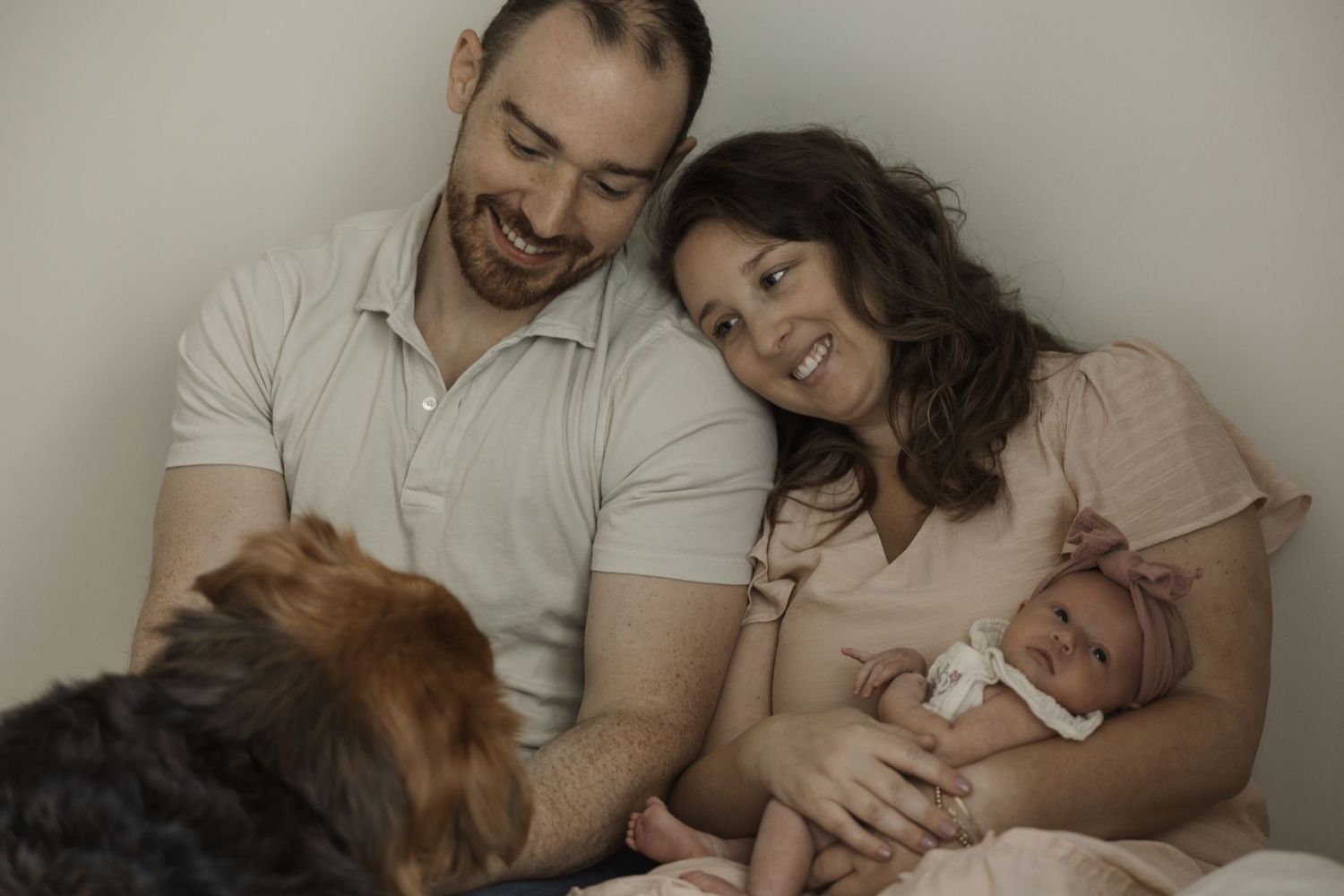 Smiling parents cuddle their newborn baby with a dog in a warm family portrait.