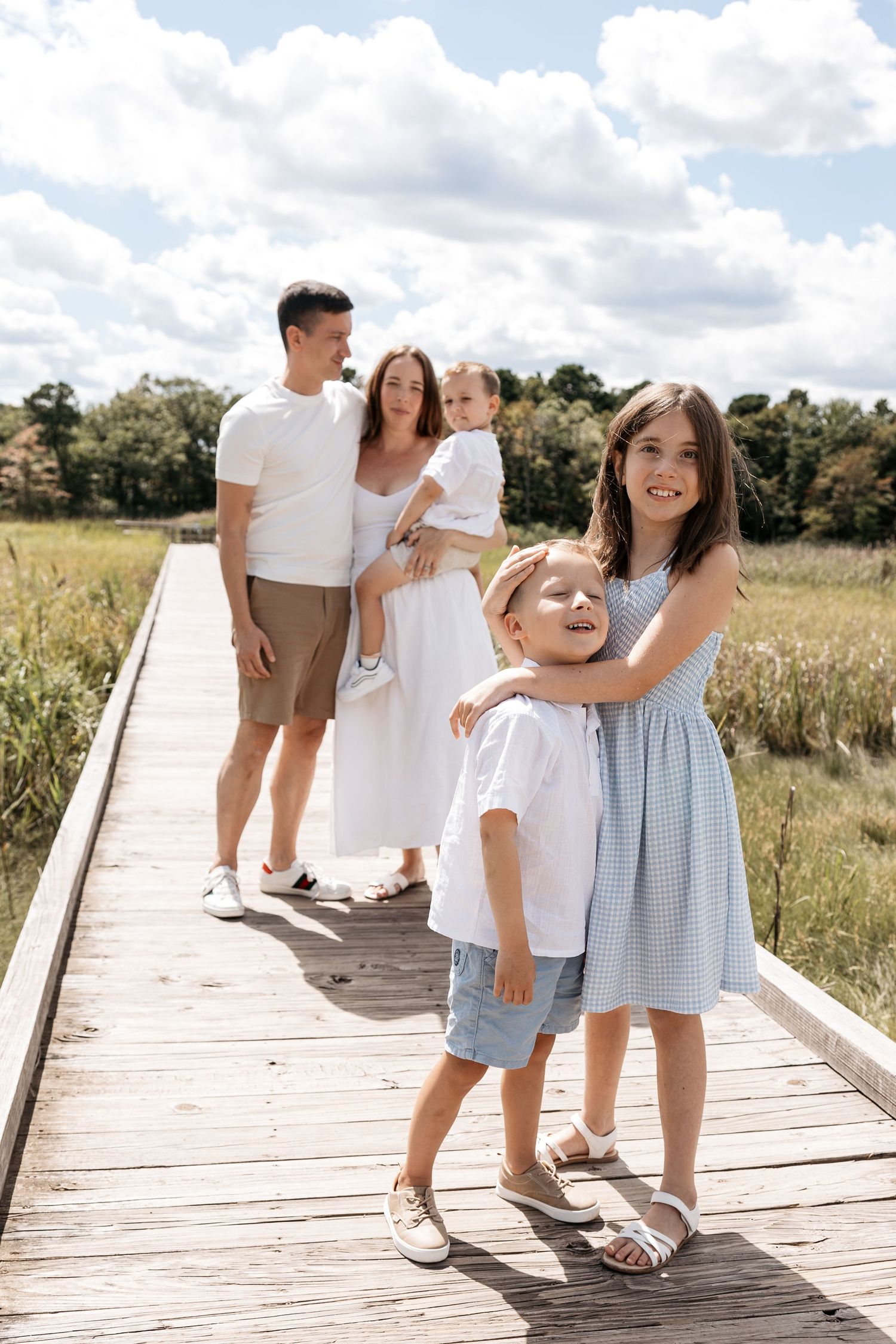 A family of five poses on a wooden boardwalk surrounded by green fields on a sunny day.