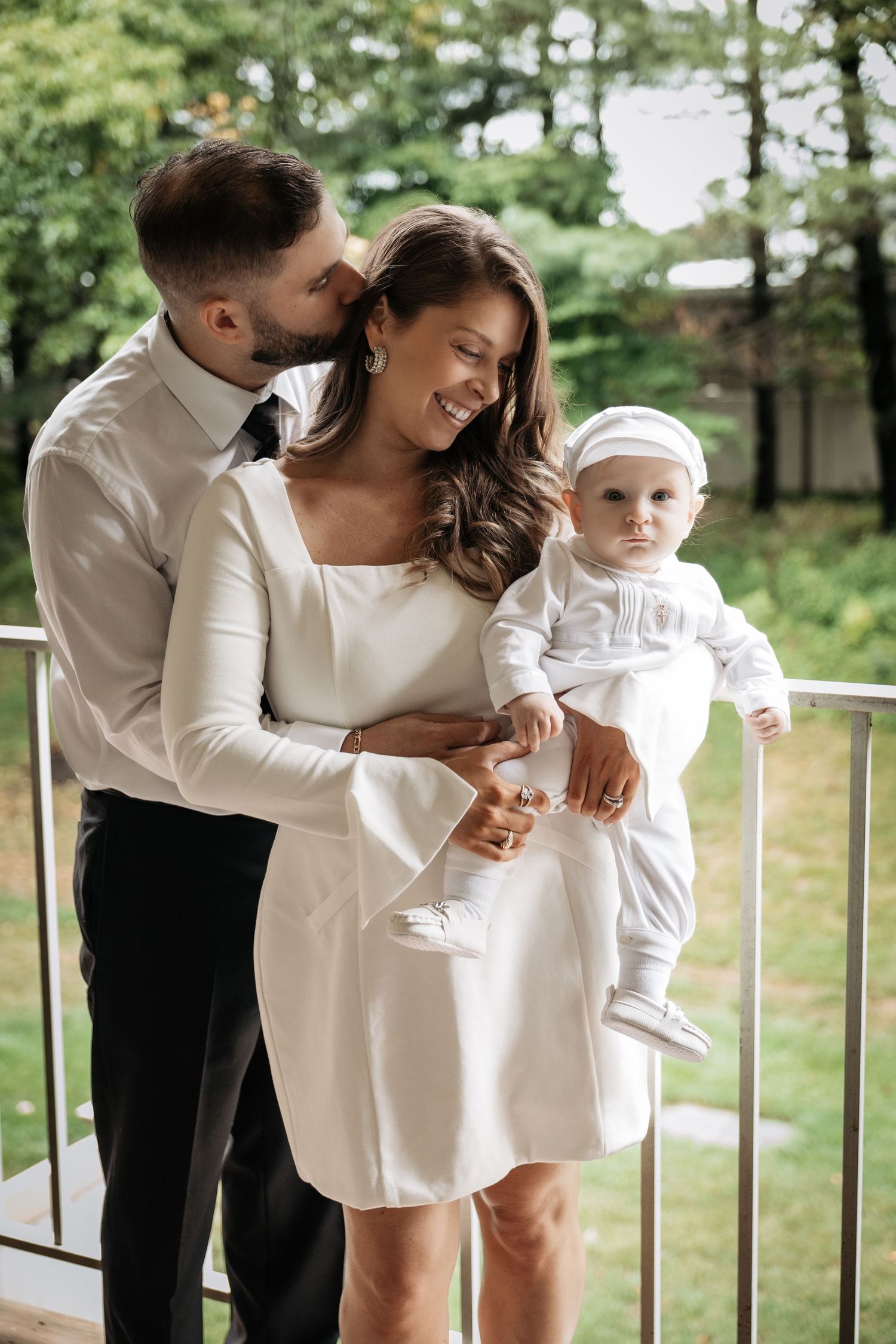 Loving father kisses mother's cheek while she holds baby in white christening outfit on outdoor balcony.