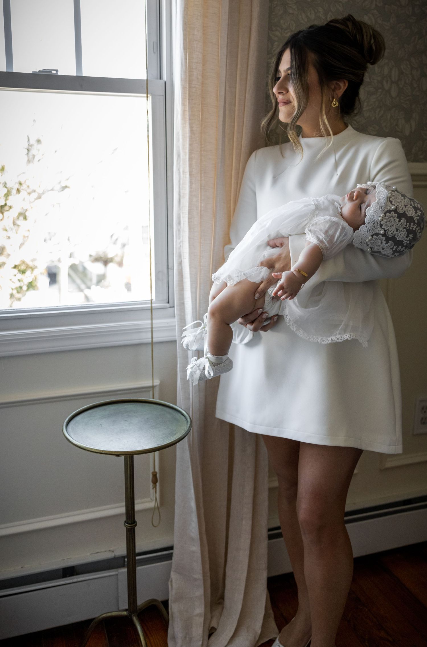 Elegant mother in white dress holding her baby in a lace christening gown by a sunlit window.