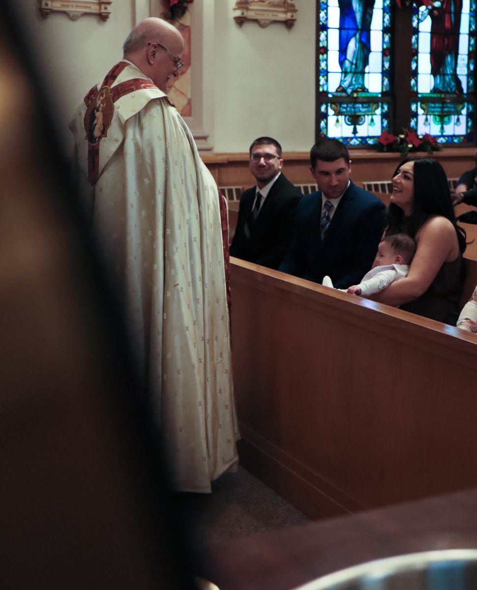 Priest in white vestments greets family seated in church pews during a baptism ceremony.