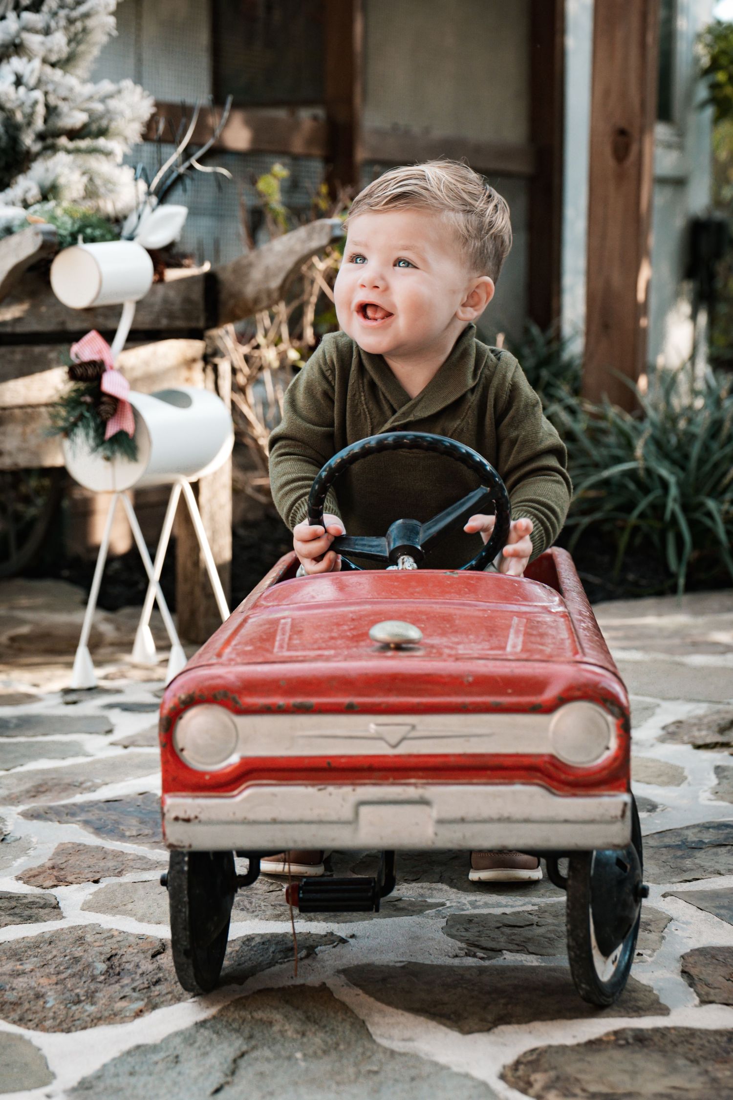 Happy toddler boy in olive hoodie smiling while sitting in a vintage red pedal car outdoors.